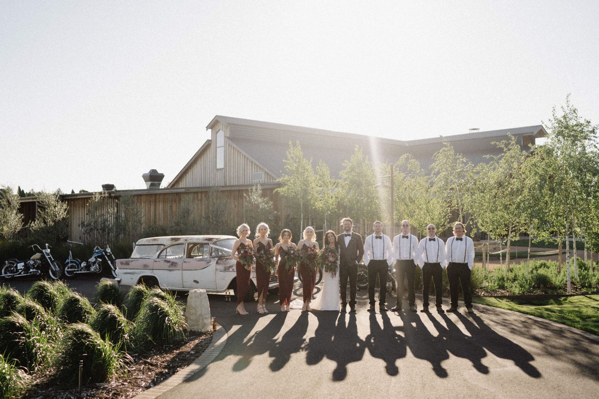 Wedding party posing in front of a rustic building with a vintage truck. Sun shines, casting long shadows.