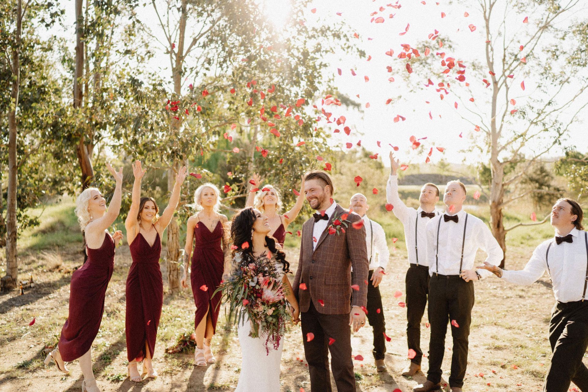 Wedding party throwing rose petals, bride and groom smiling, sunny outdoor setting.