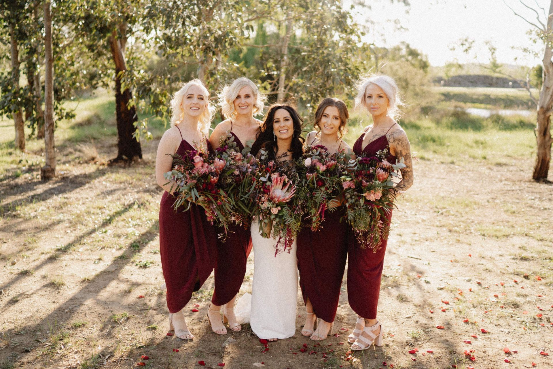 Bride and bridesmaids in burgundy dresses, holding bouquets, smiling outdoors.