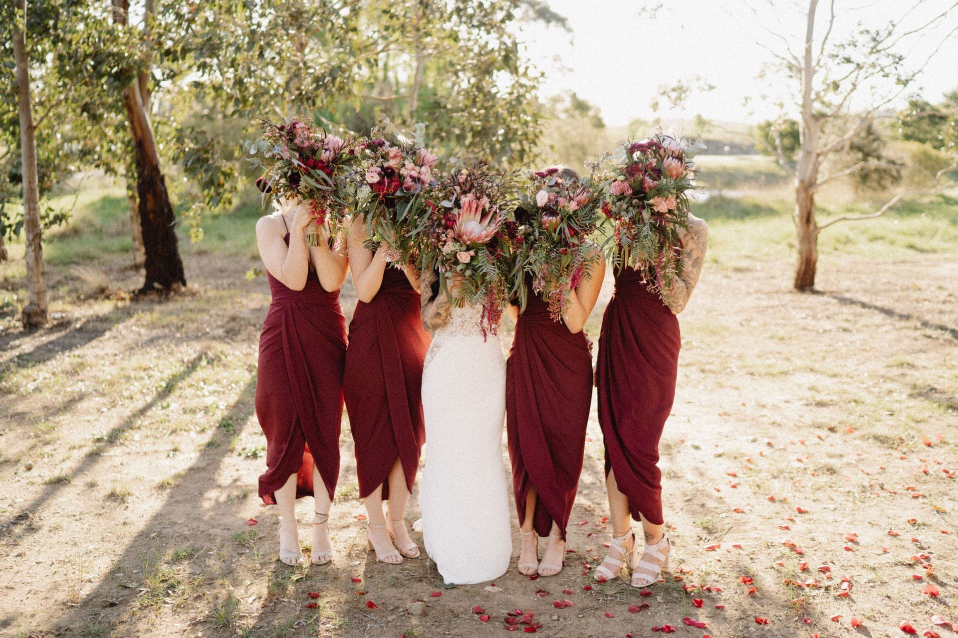 Bride in white gown surrounded by bridesmaids in burgundy dresses, holding large bouquets, standing outside.
