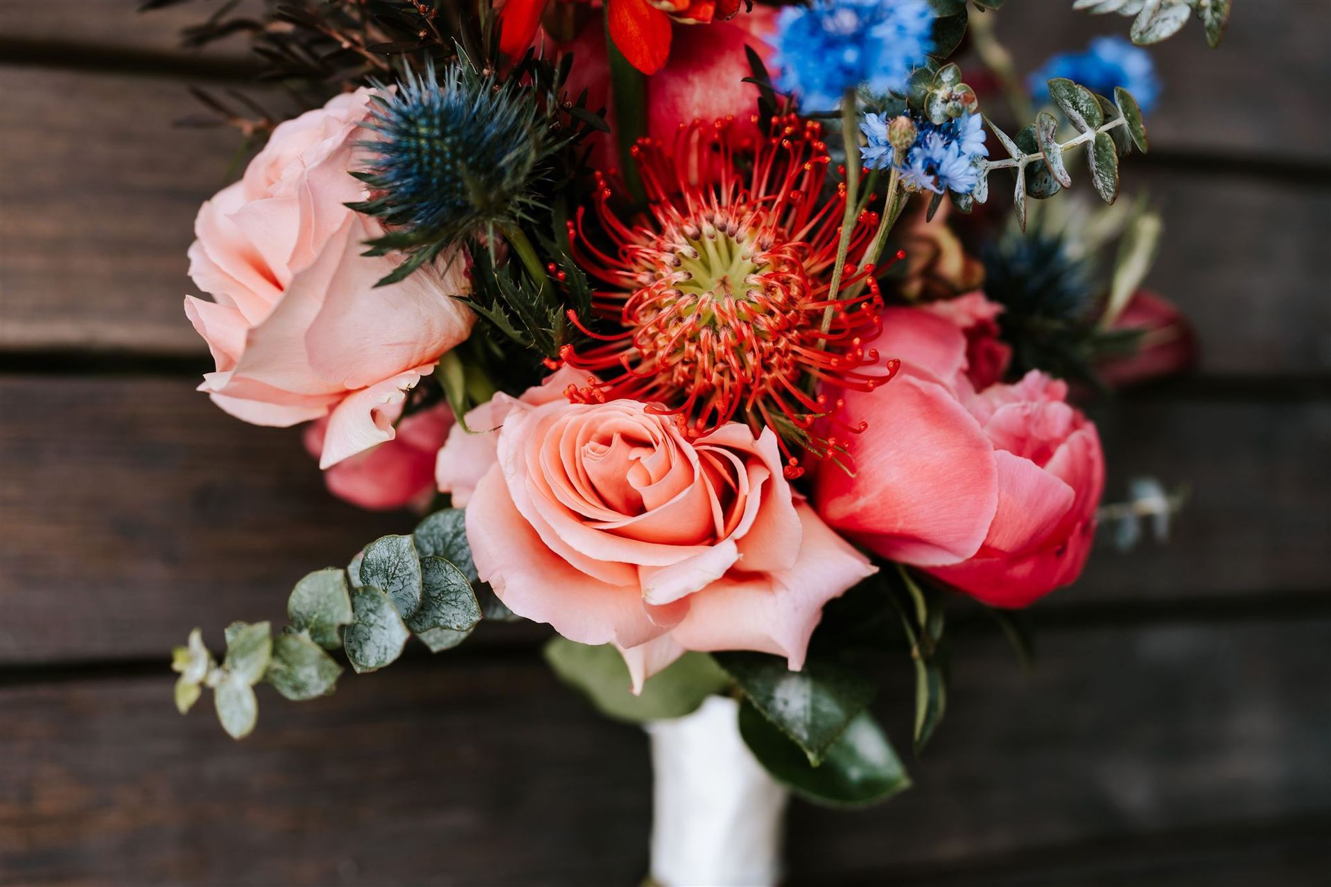 Bouquet of pink, red, and blue flowers, including roses and thistles, wrapped in white ribbon against a wood background.