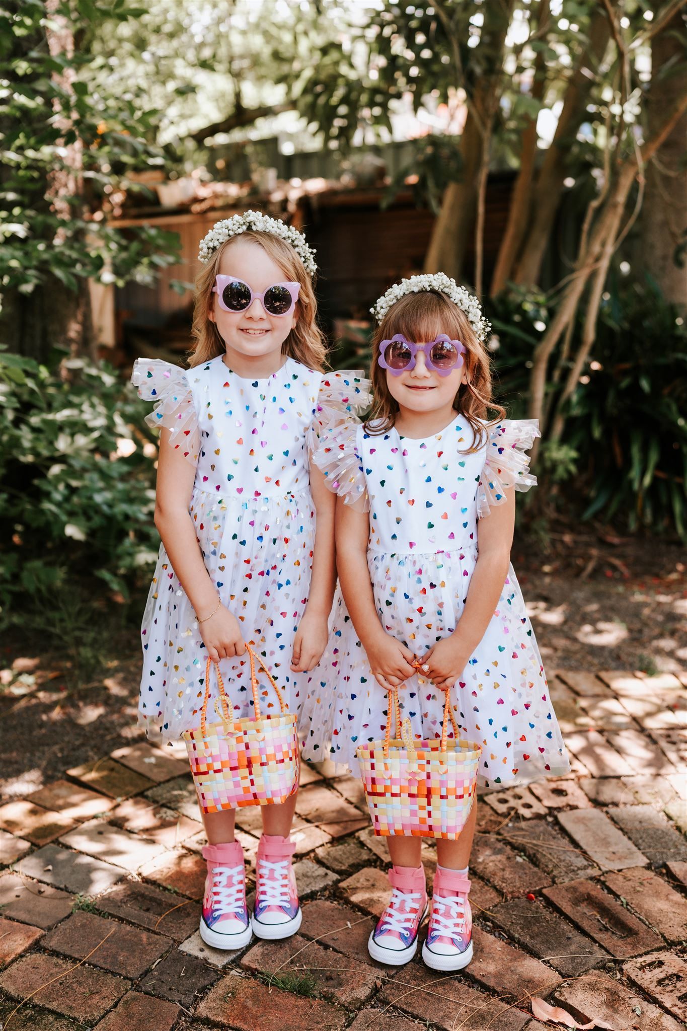 Two girls in white polka dot dresses with heart sunglasses and flower crowns, holding baskets.