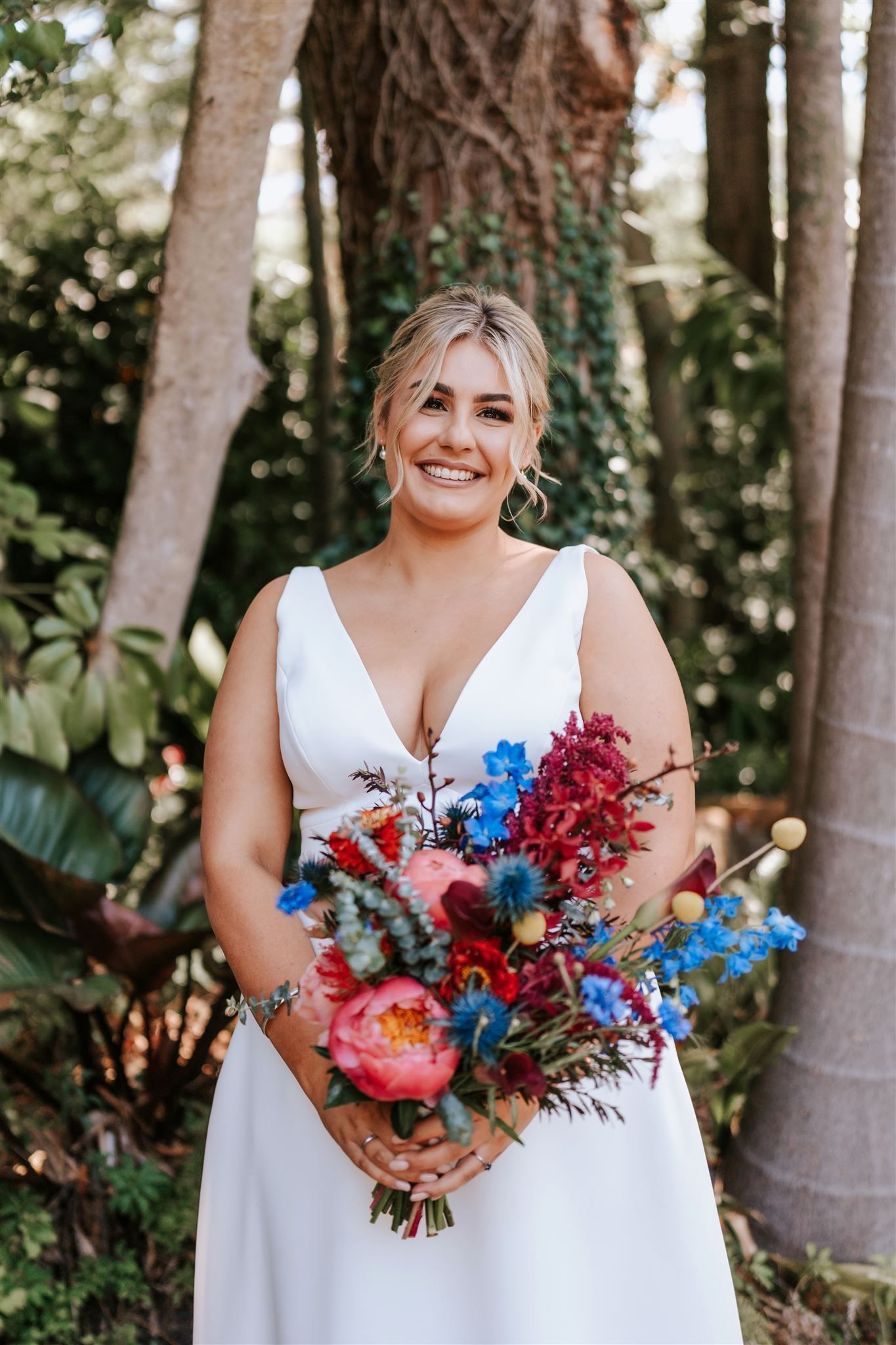 Bride in white dress holding colorful bouquet, smiling outdoors.