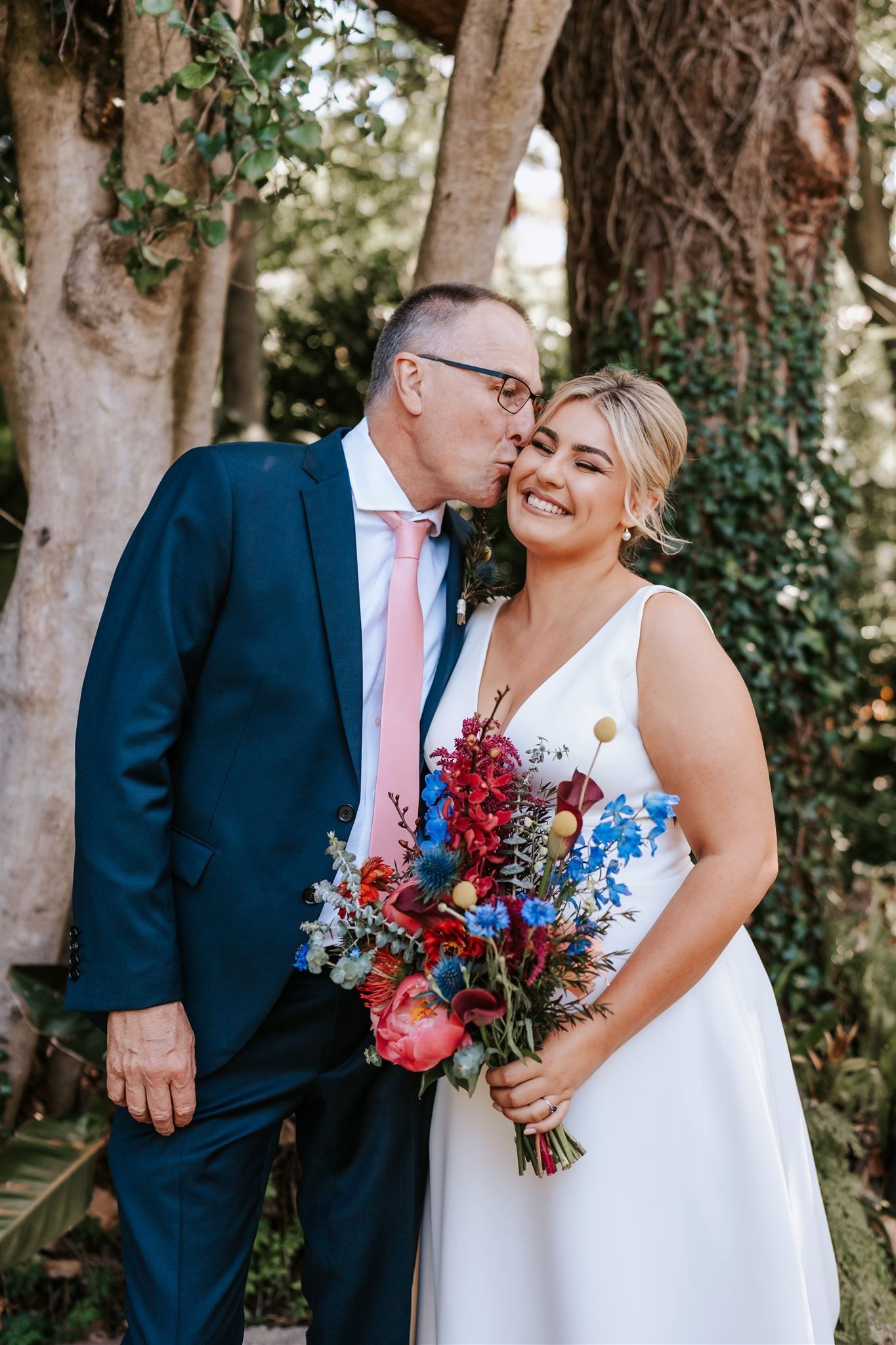 Man kissing a smiling woman holding a colorful bouquet, standing near a tree, likely at a wedding.