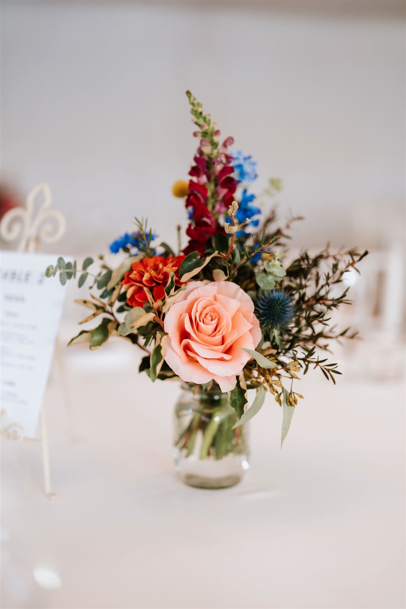 Floral arrangement in a glass jar with a peach rose, red, blue, and green blooms. Table setting.