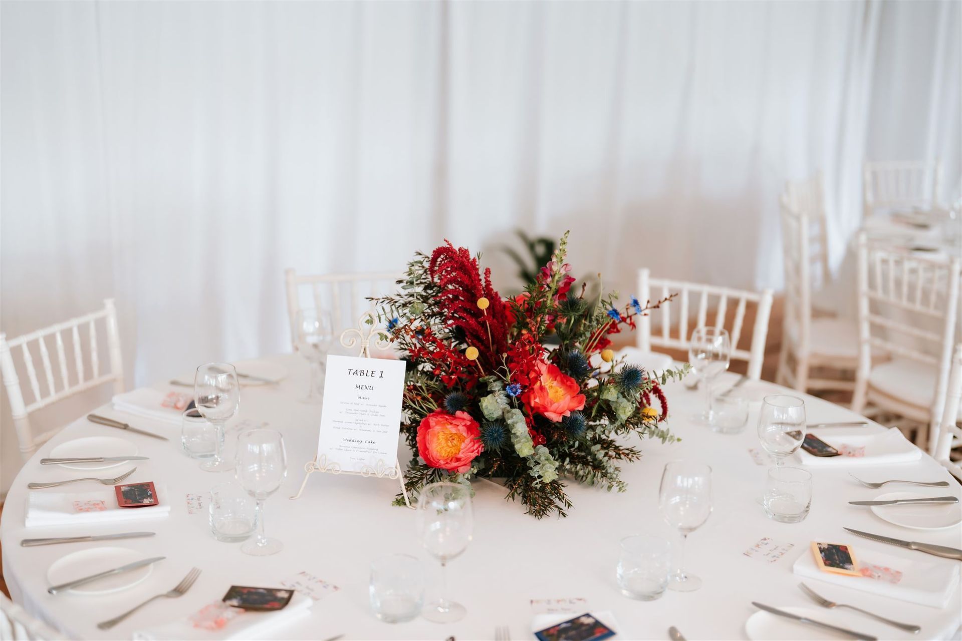 Round table set for a wedding, with floral centerpiece and place settings. White chairs and backdrop.