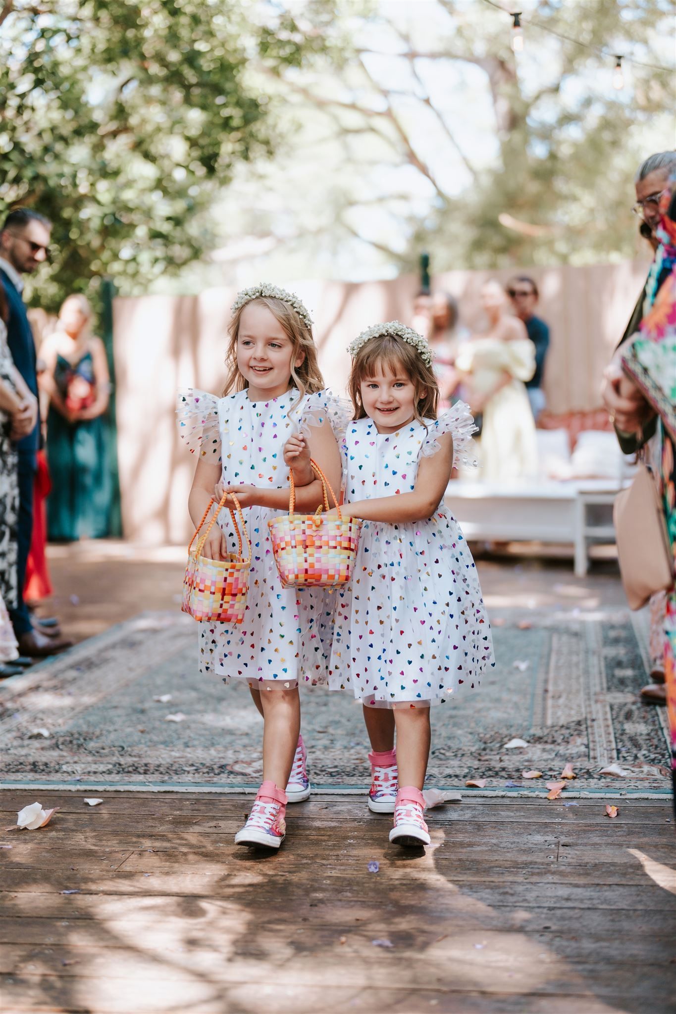 Two young girls in polka-dot dresses walk down an aisle, carrying baskets, at an outdoor wedding.