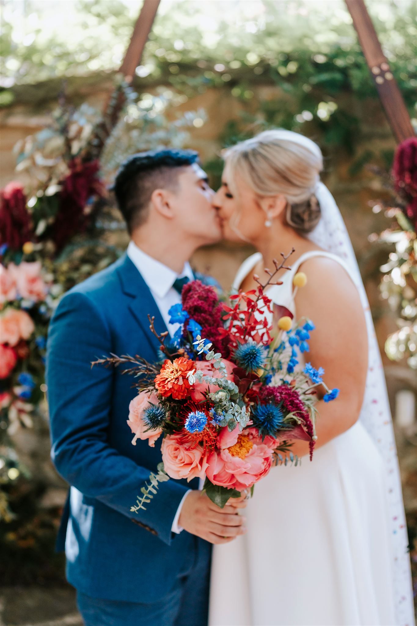 Couple kissing at wedding, vibrant floral bouquet. Blue suit, white dress. Ceremony arch in background.