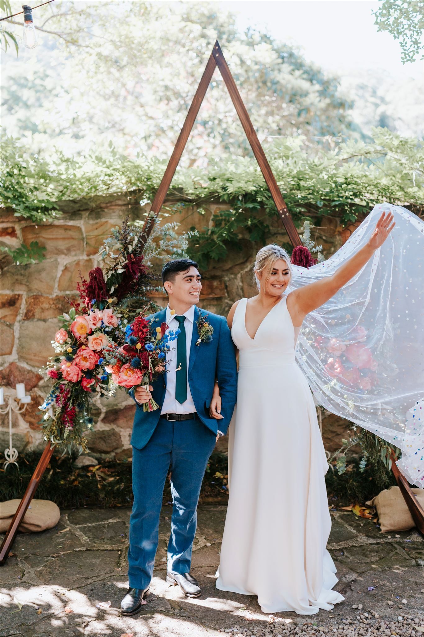 Newlyweds in front of a floral arch; the bride raises veil, groom smiles holding bouquet, blue suit, white dress.