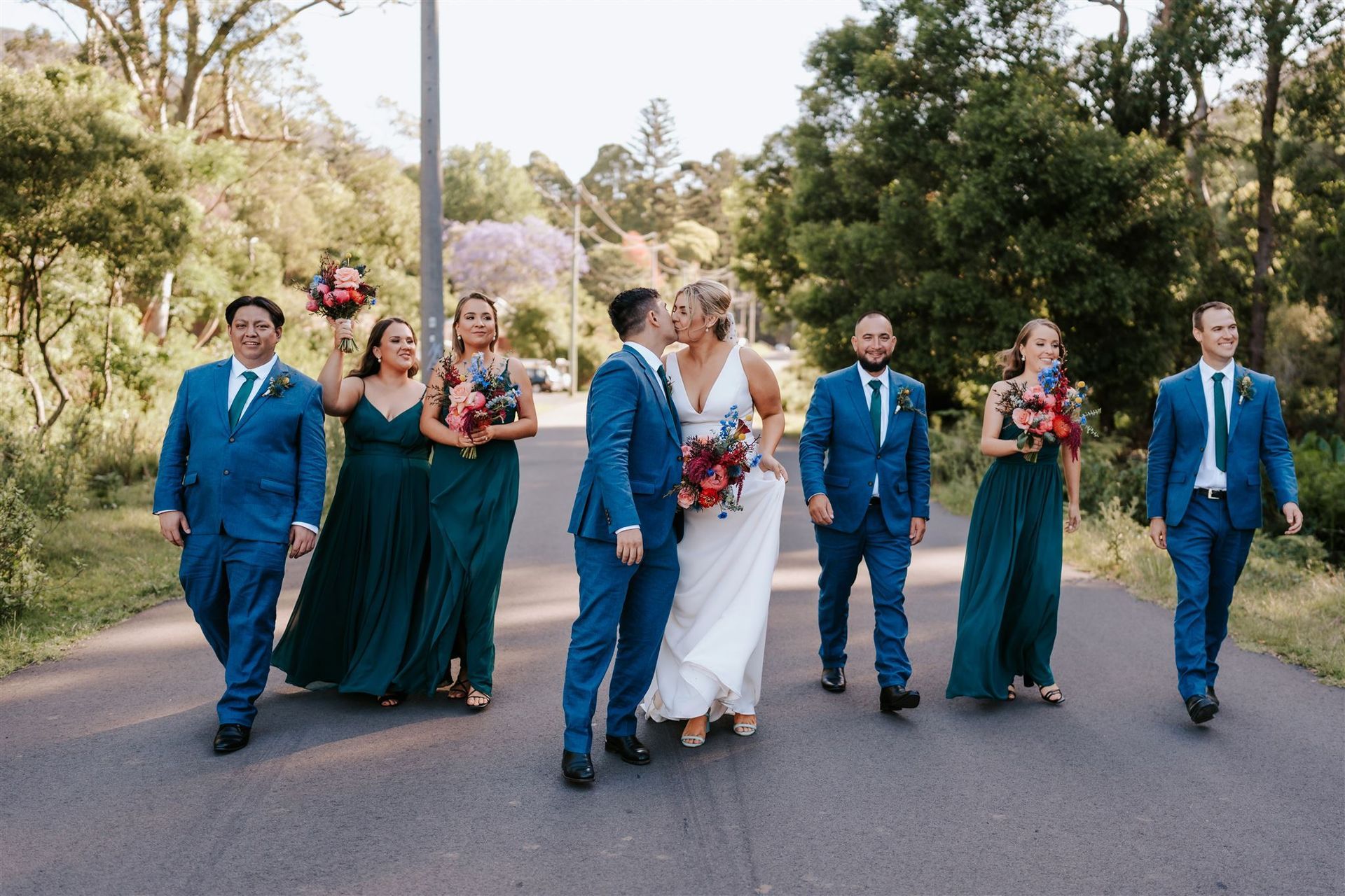Wedding party on a paved path in a park. The couple kisses, surrounded by bridesmaids in emerald and groomsmen in blue.