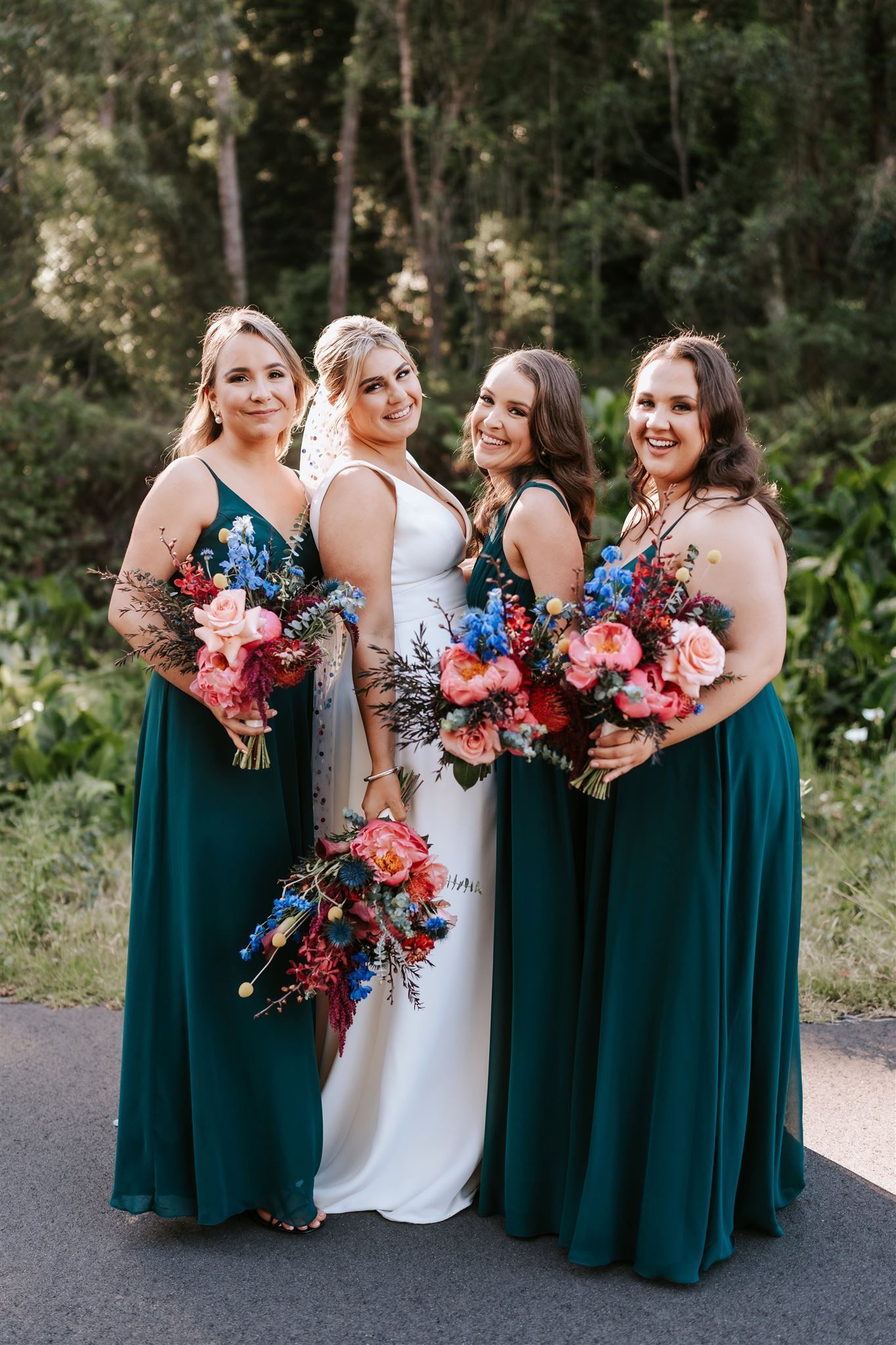 Bride and bridesmaids posing outdoors. Bride in white dress, bridesmaids in teal, holding bouquets.