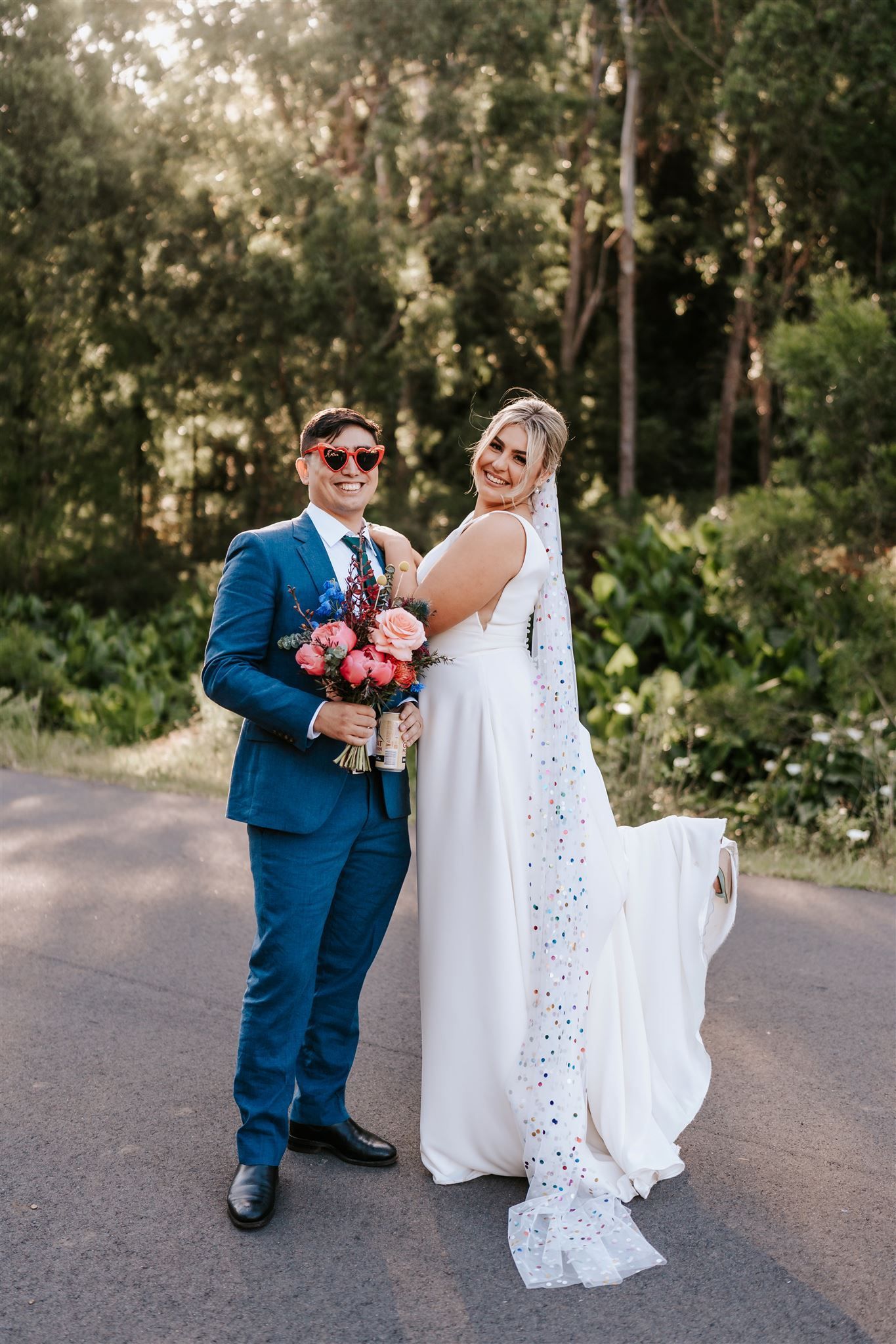 Newlyweds pose outdoors; groom in blue suit and sunglasses, bride in white dress and veil holding flowers.