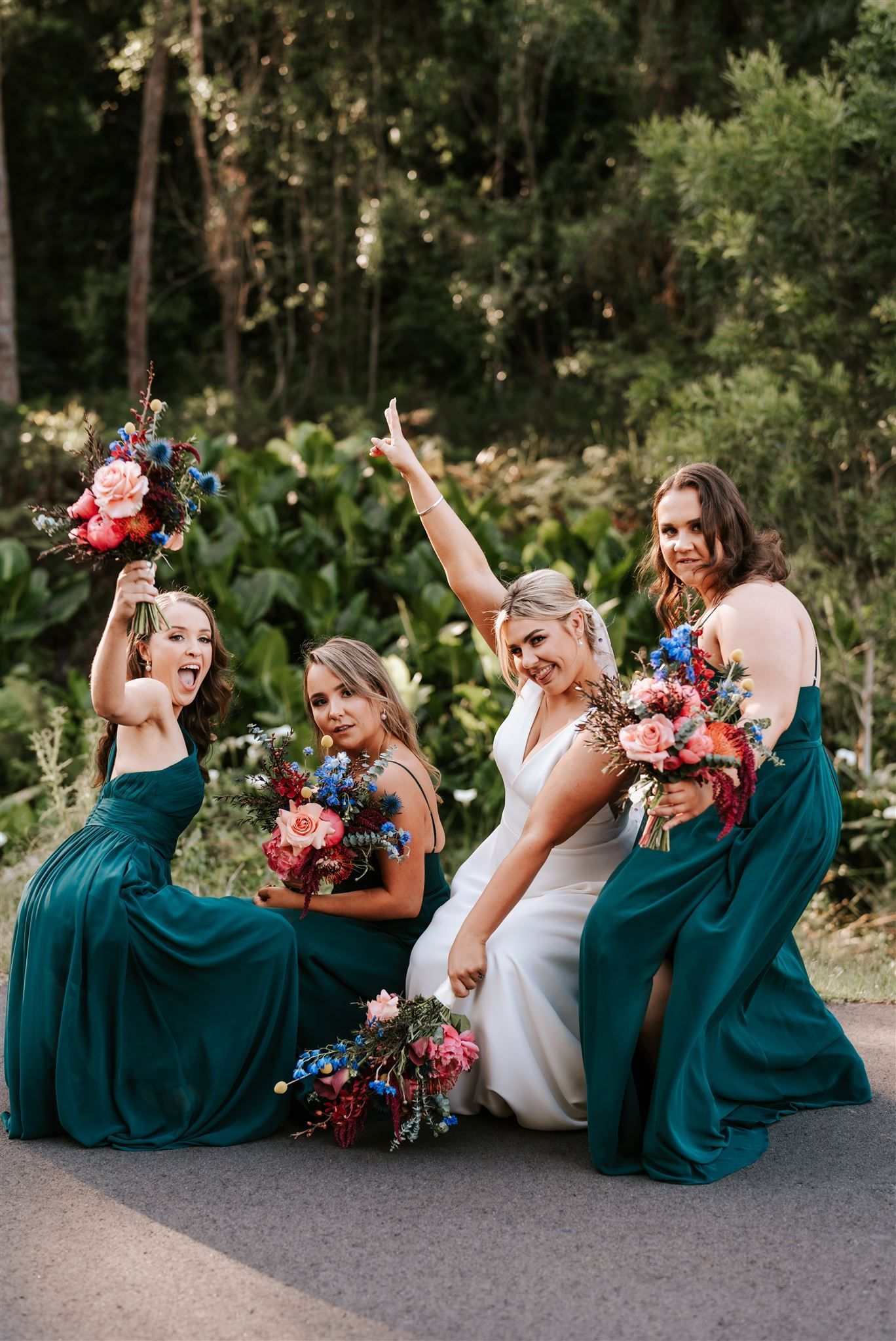 Bride with bridesmaids in teal dresses, celebrating with bouquets. Outdoors, forest backdrop.