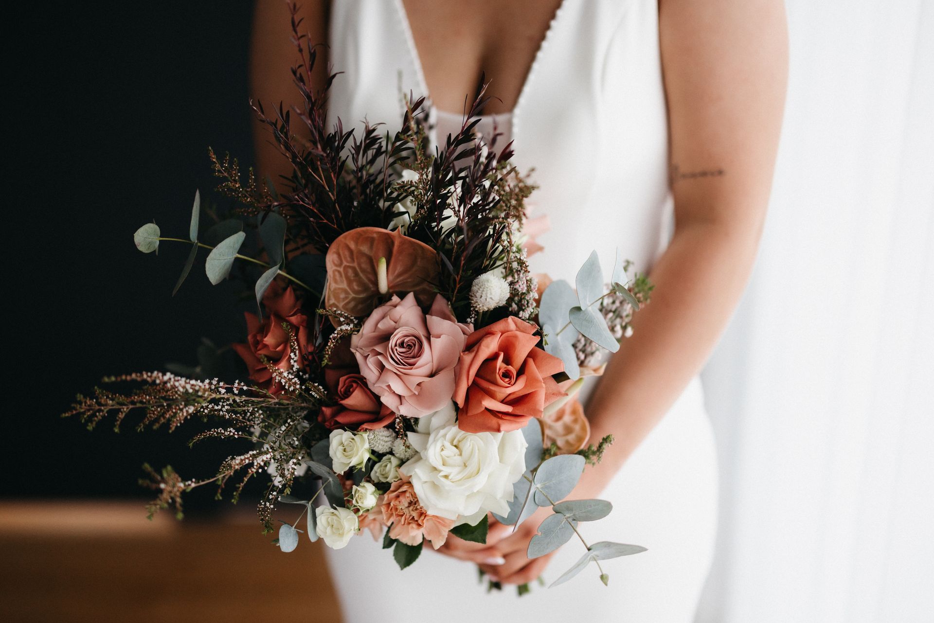 Bride holding a bouquet of red, pink, and white flowers against a white dress and dark wall.