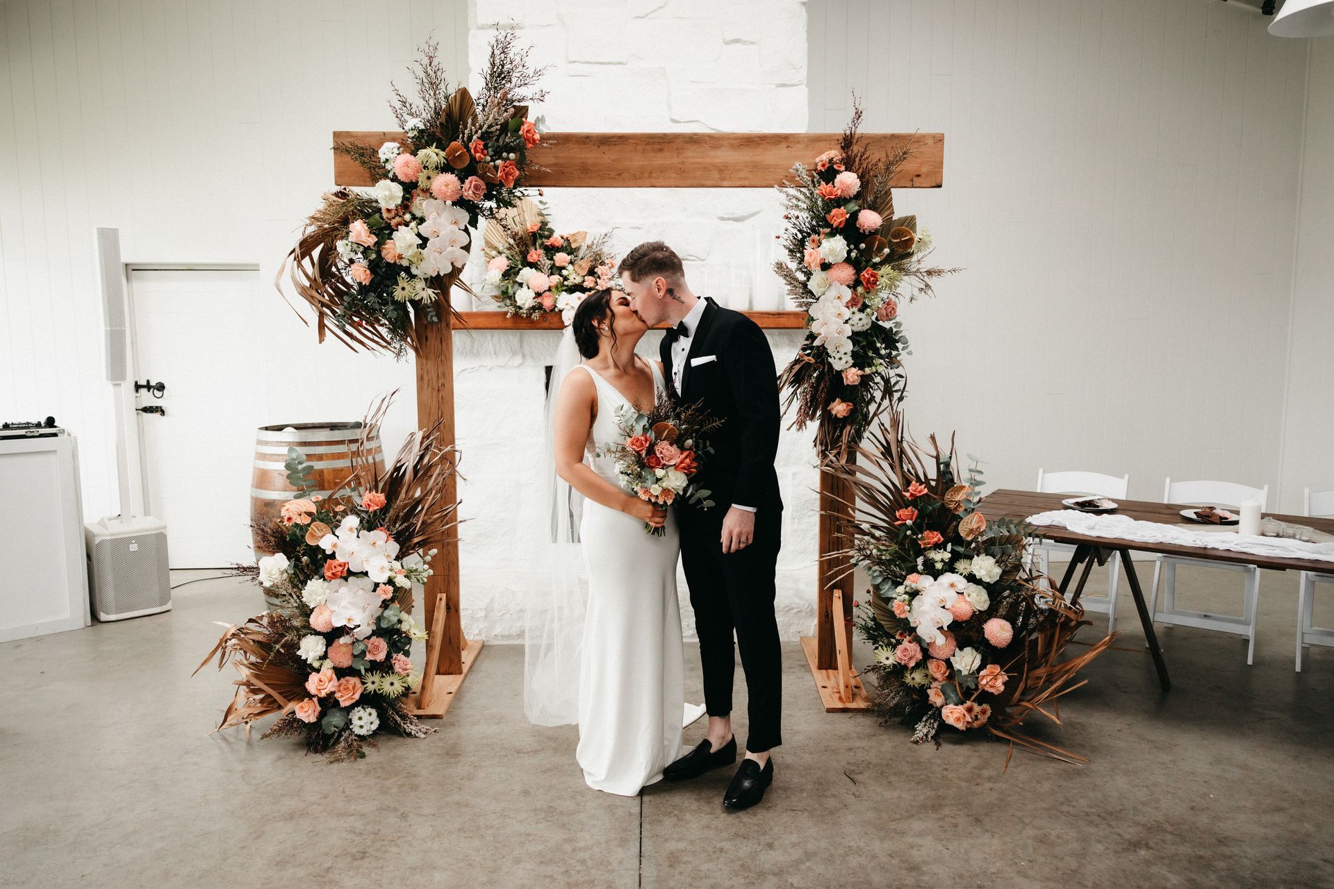 Newlyweds kissing under floral archway; inside, neutral tones, warm colors.