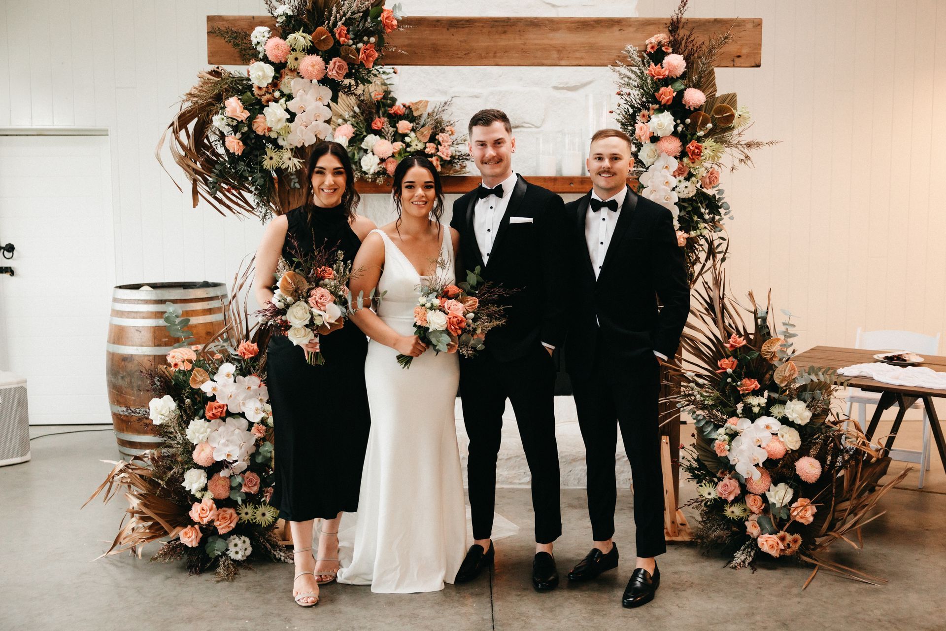 Wedding party posing in front of floral archway; bride and bridesmaids, groomsmen in black suits.