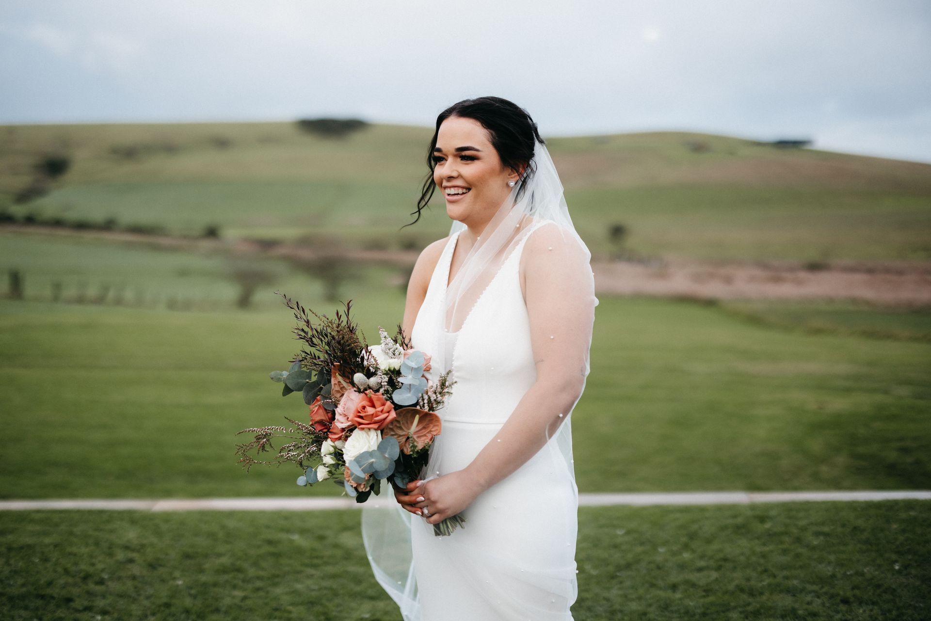 Bride laughing outdoors, holding bouquet. She wears a white dress and veil in a field with green hills.