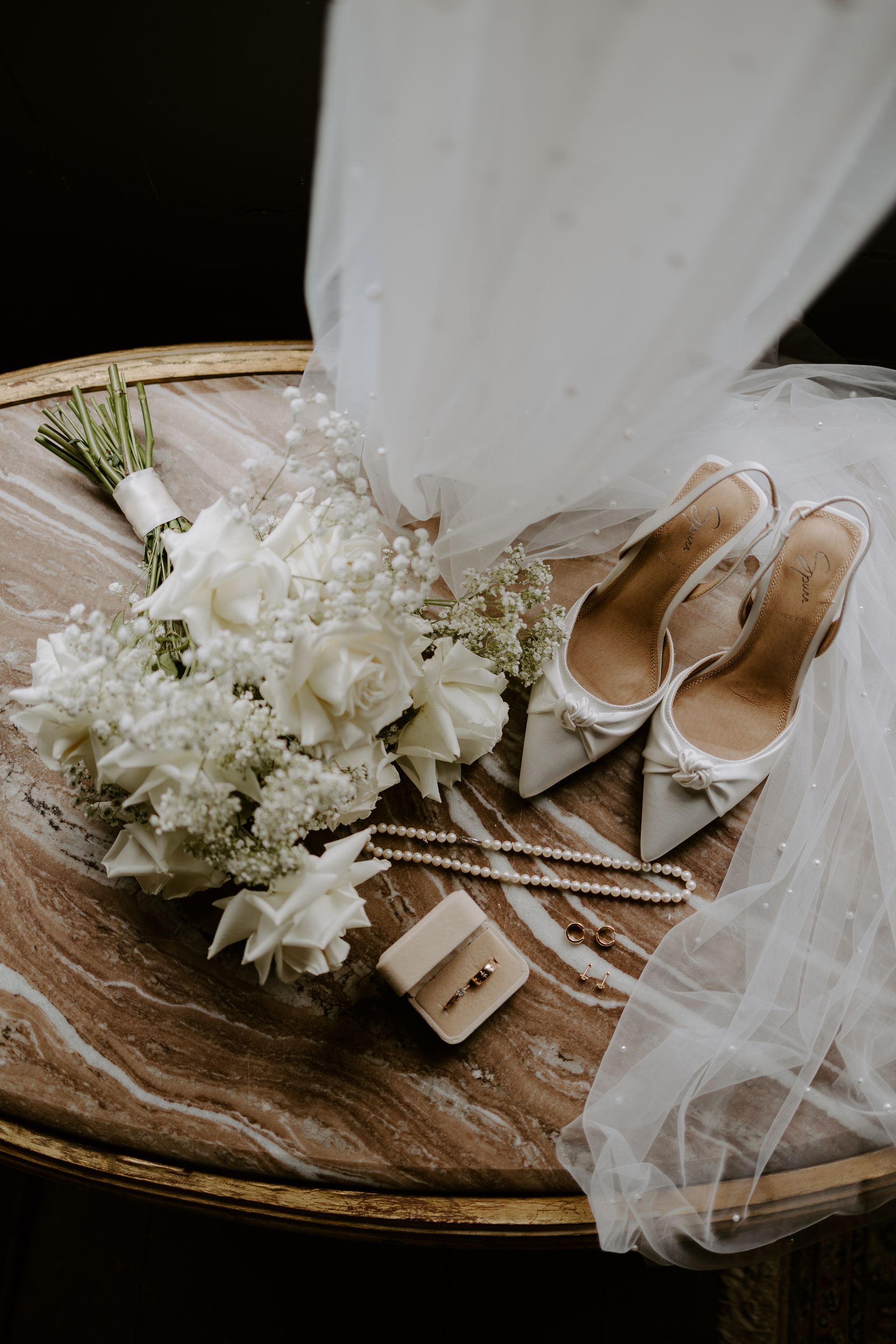 Wedding details: bouquet, shoes, veil, rings, and jewelry on a marble table.