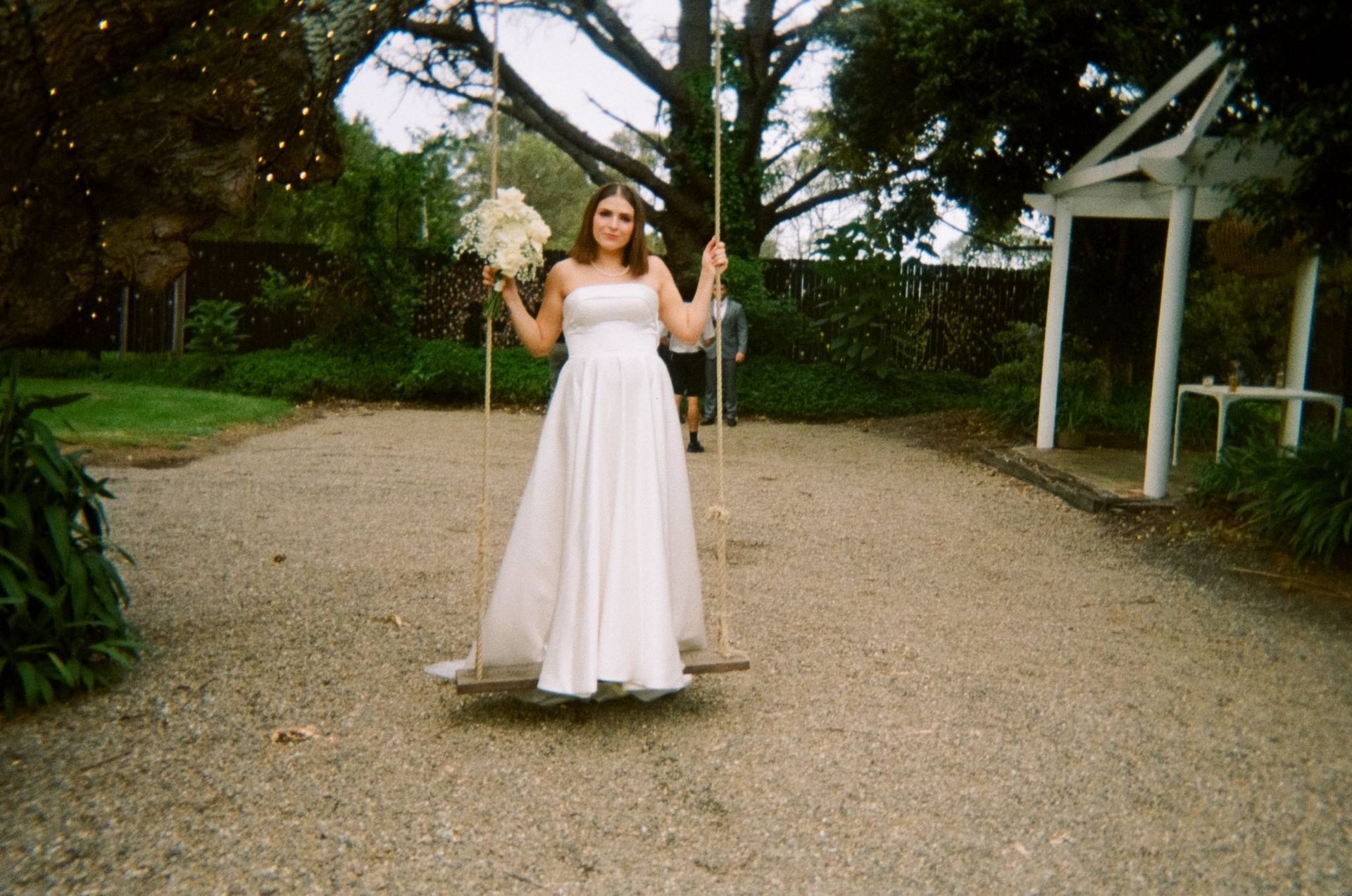 Bride in a white dress on a swing outdoors, holding flowers. Gravel ground, greenery, and small white structure.