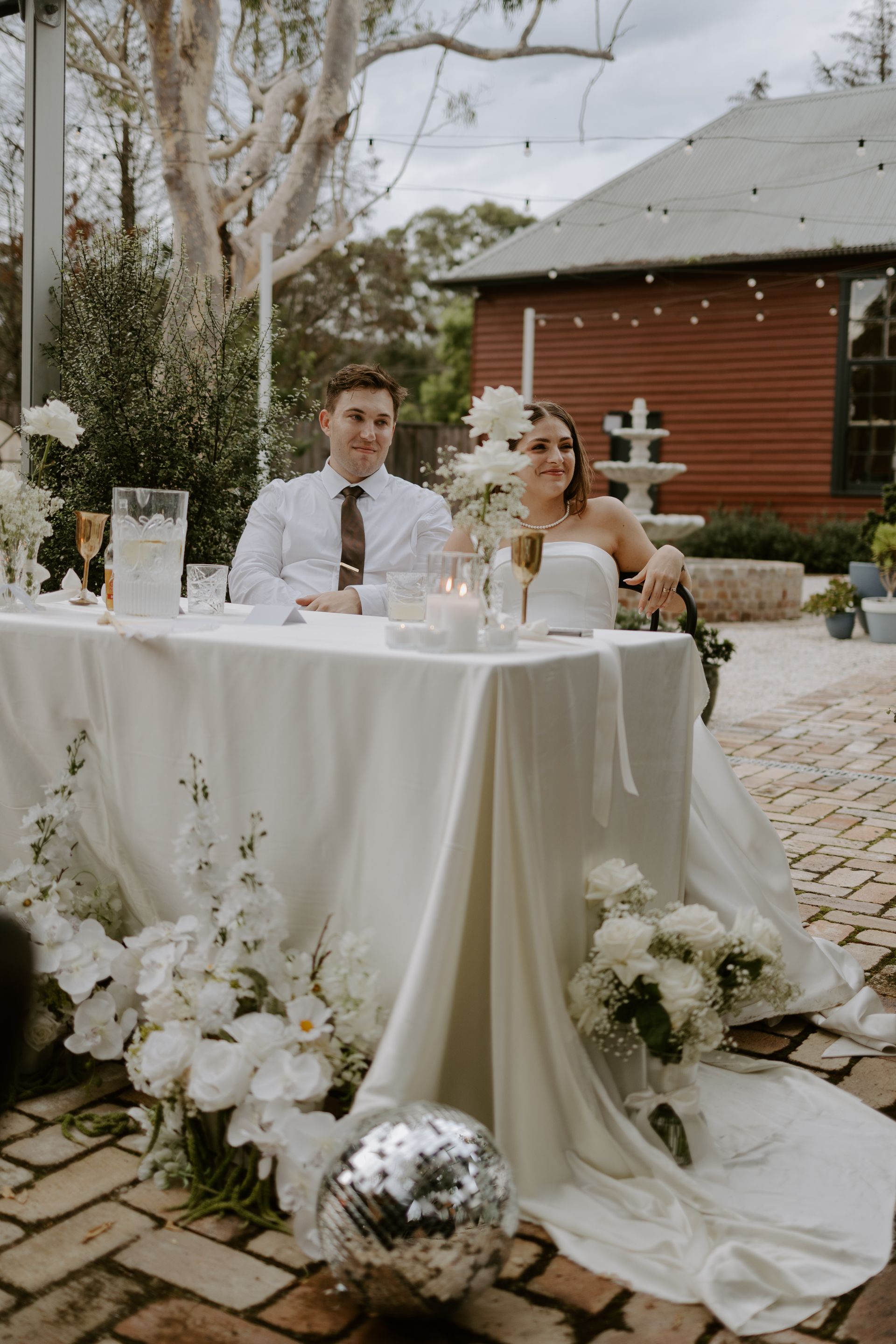 Newlyweds at a wedding reception table decorated with white flowers and a disco ball. Outdoor setting.