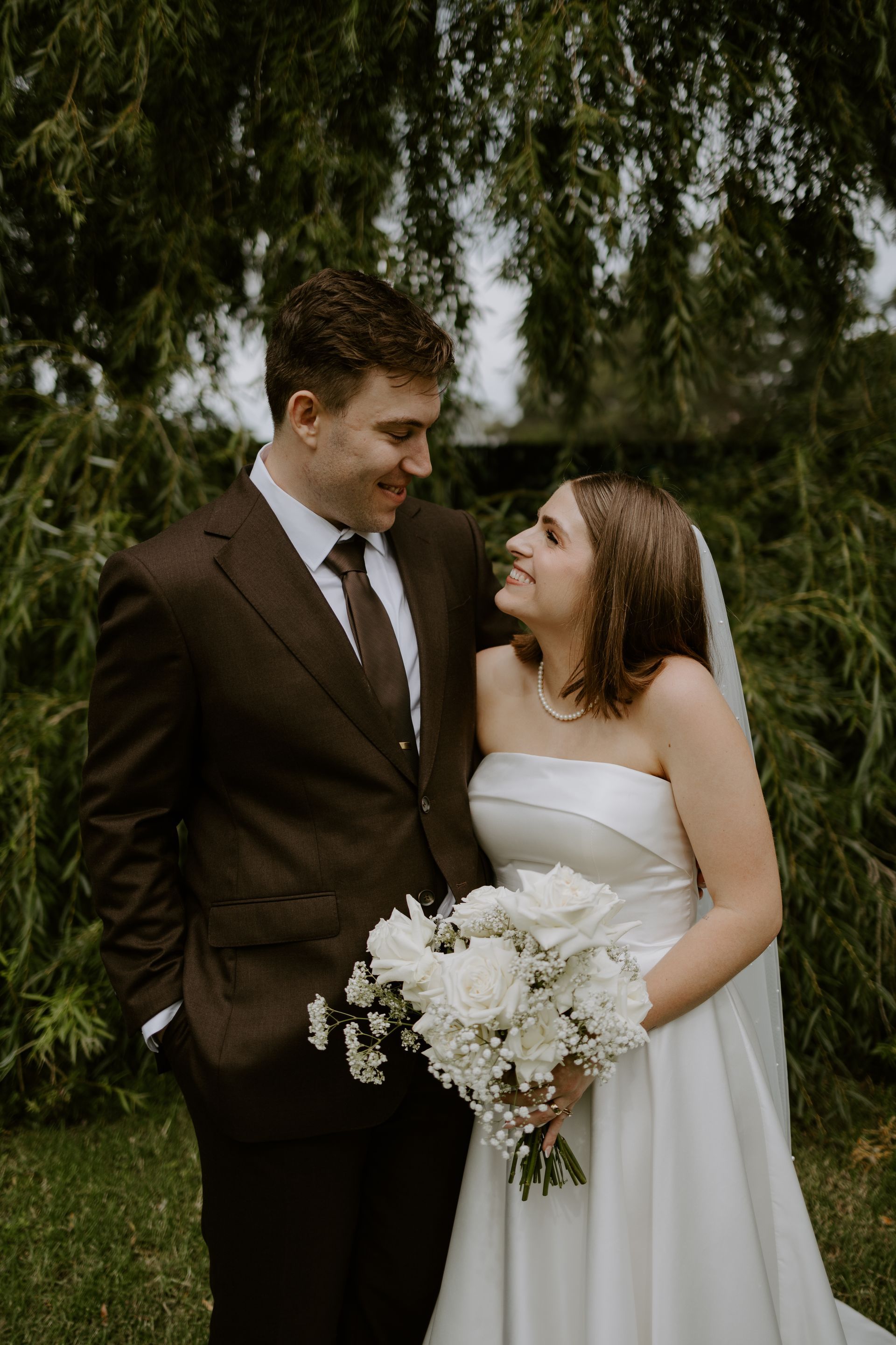 Bride and groom smiling at each other, holding bouquet, under weeping willow tree.
