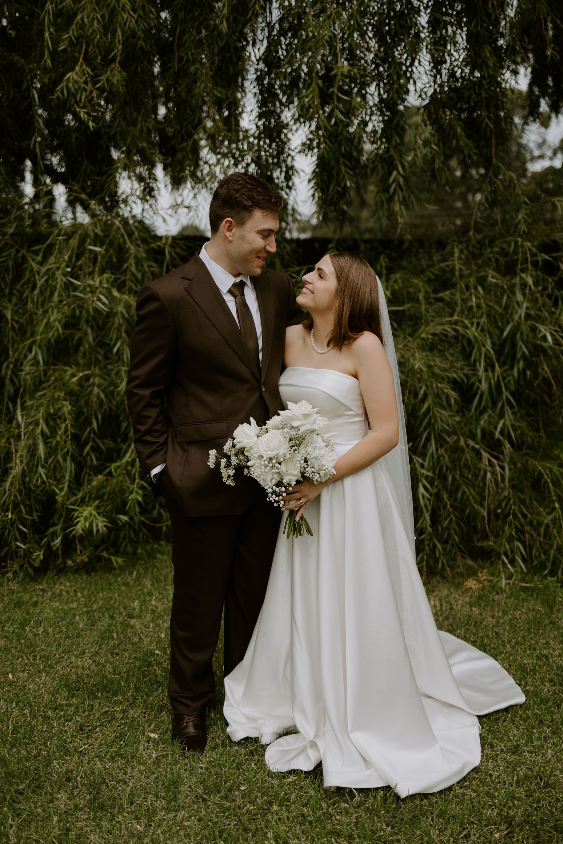 Wedding couple smiling at each other in front of a weeping willow tree. The bride holds a bouquet.