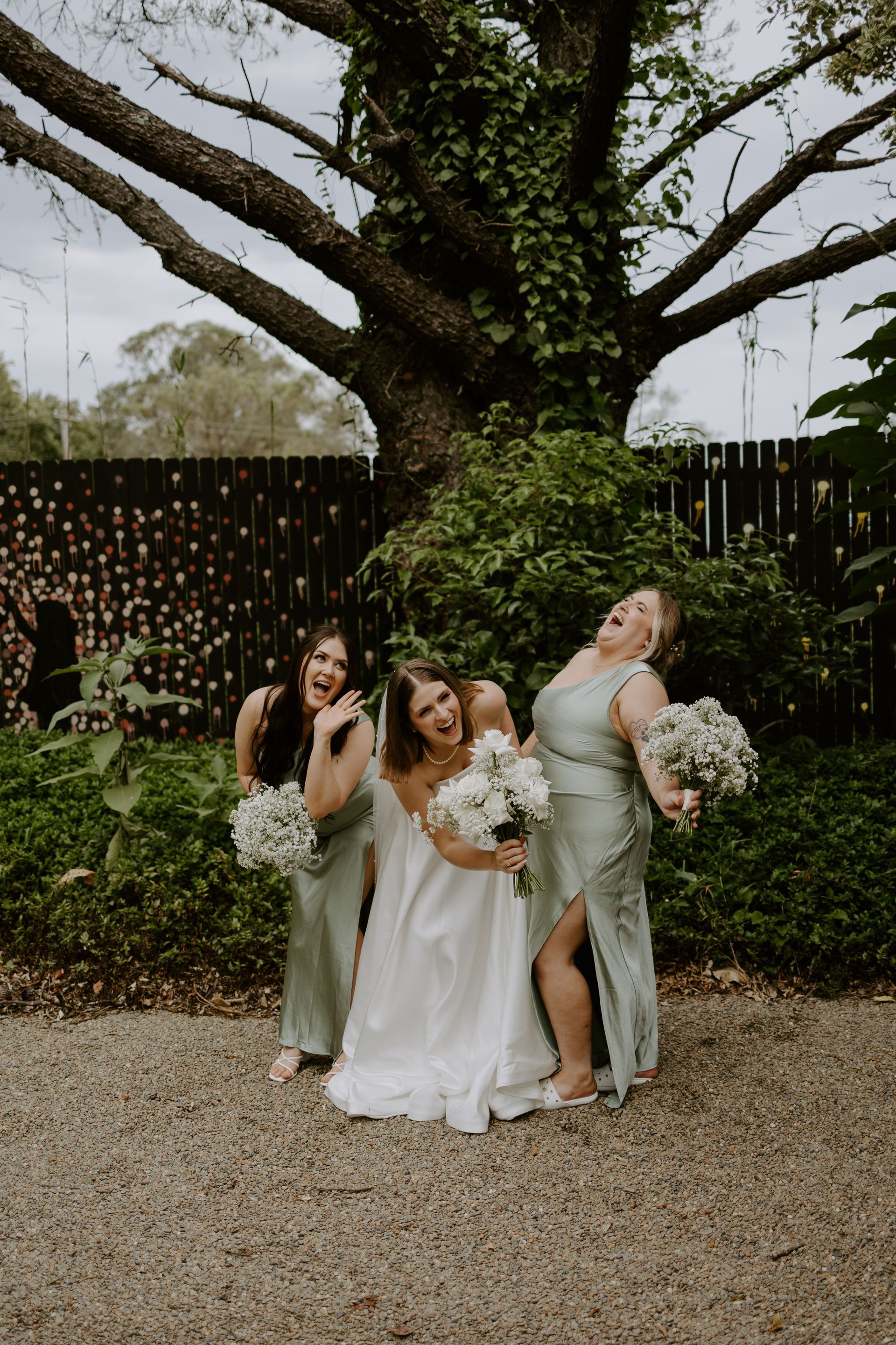 Bride and bridesmaids laughing, holding bouquets in garden setting with large tree and fence.