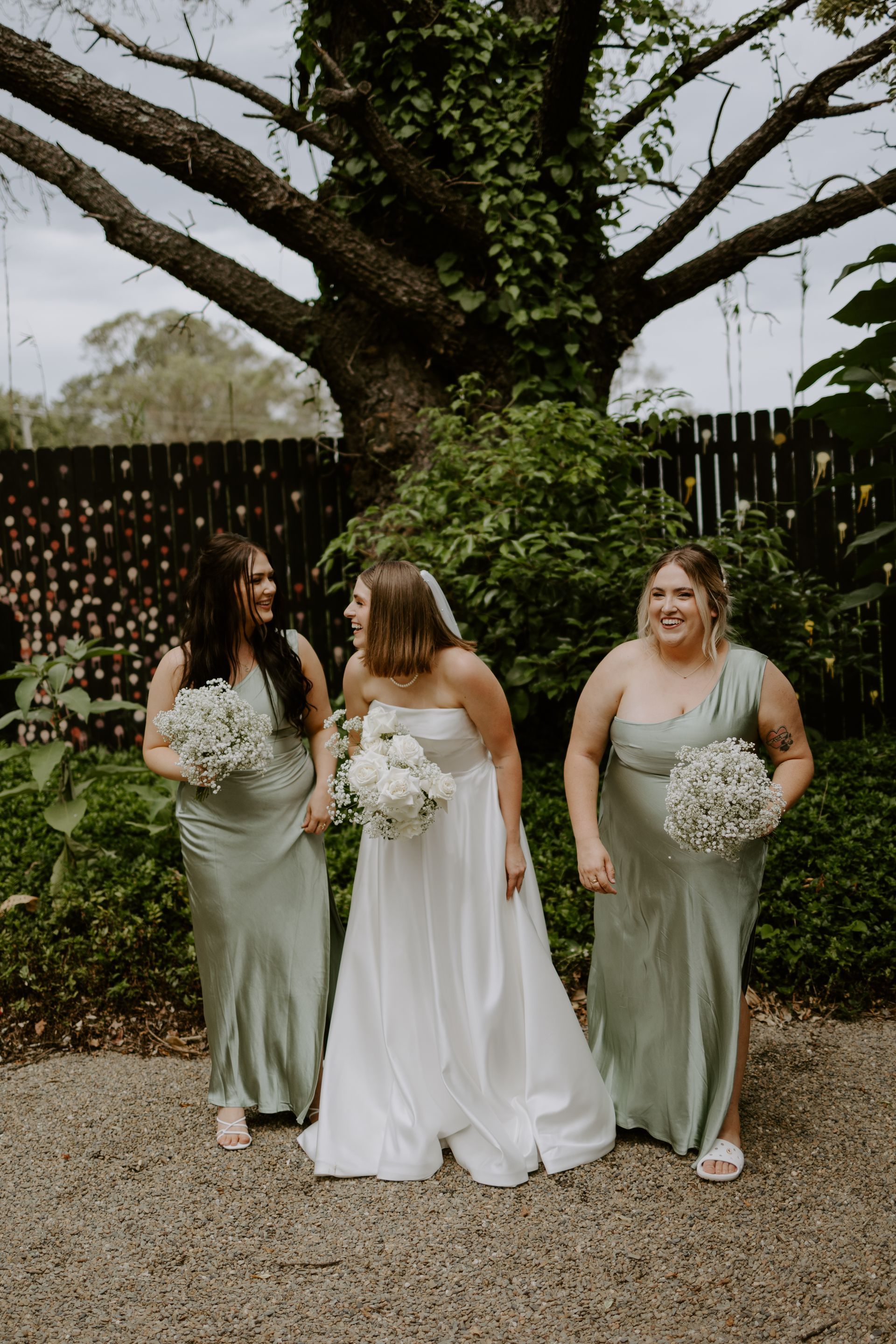 Bride and bridesmaids in sage green dresses, holding bouquets, near a tree.