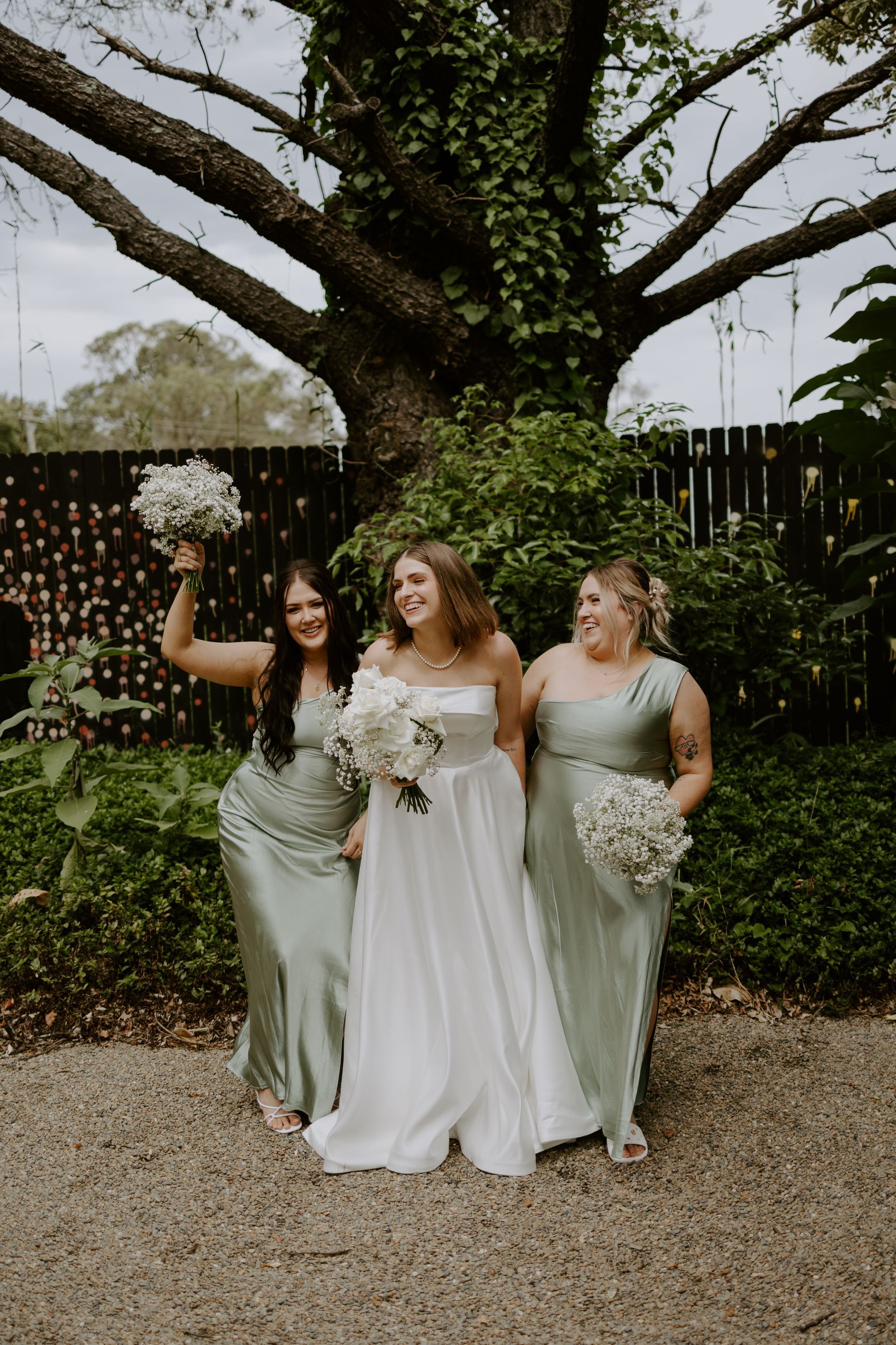 Bride and bridesmaids smiling outdoors, holding bouquets. The bride wears white, bridesmaids wear green.