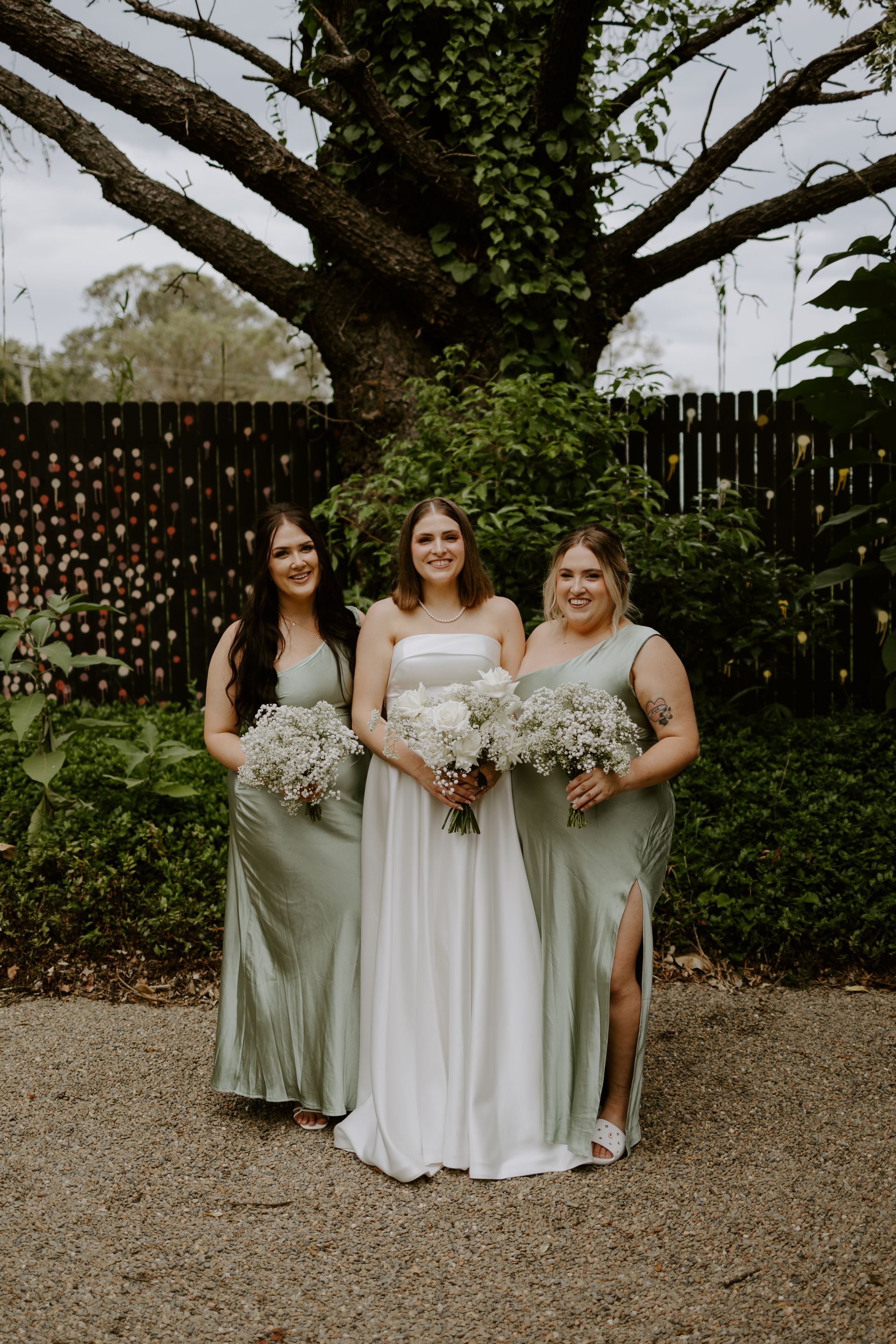 Bride and bridesmaids pose outdoors. Bride in white gown, bridesmaids in green dresses, holding bouquets.