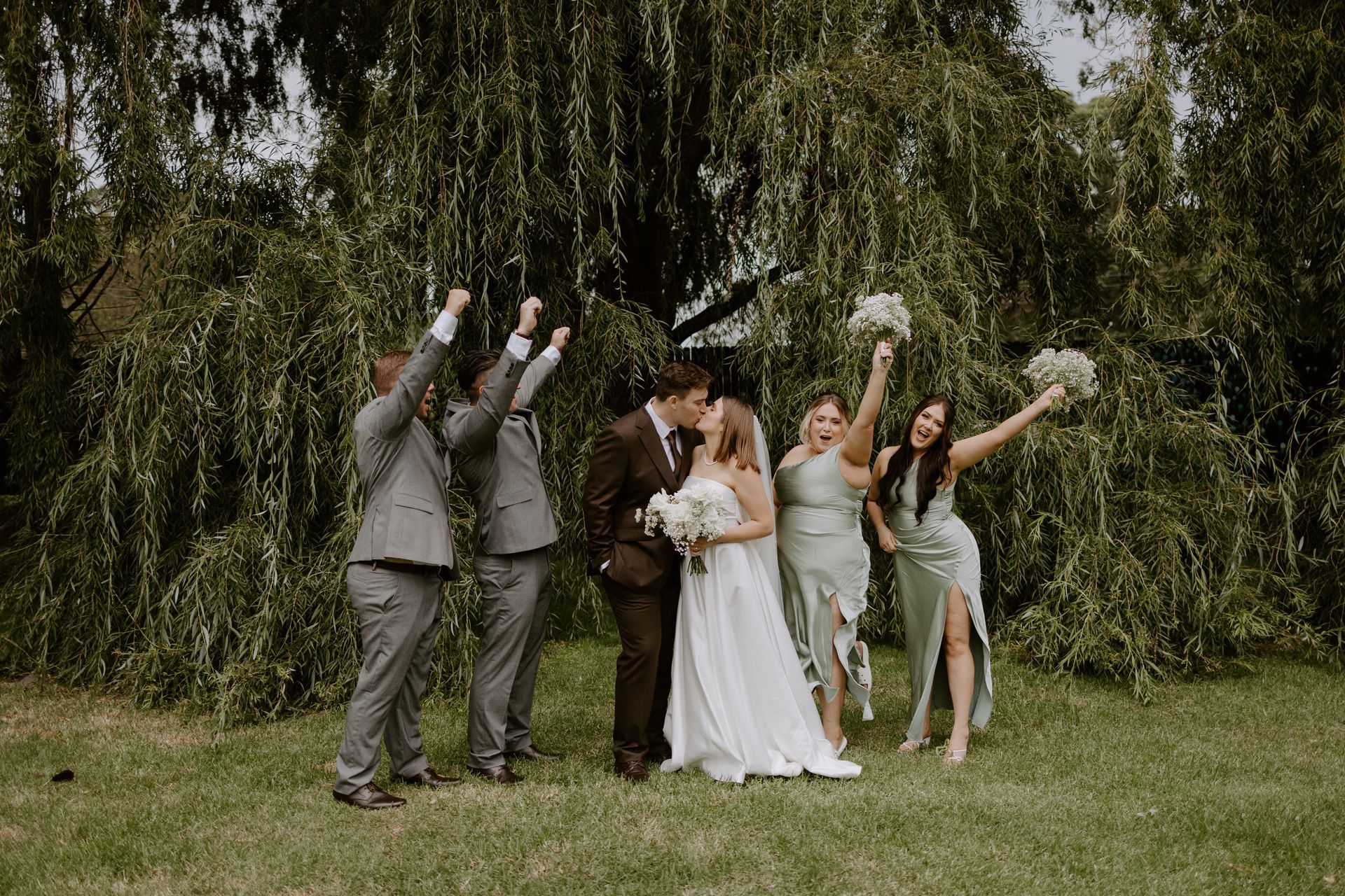 Wedding party celebrating. Couple kissing, bridesmaids holding bouquets, groomsmen raising arms, all in front of a tree.