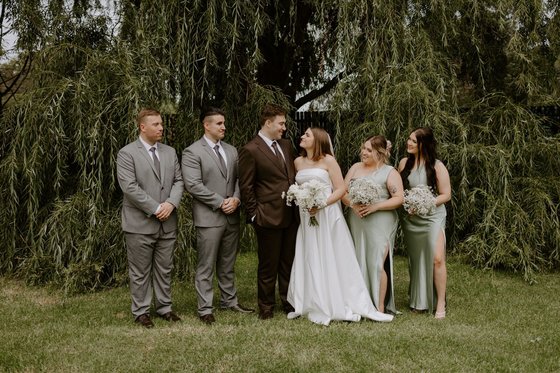 Wedding party poses under a weeping willow tree. Bride and groom face each other; attendants stand on either side.