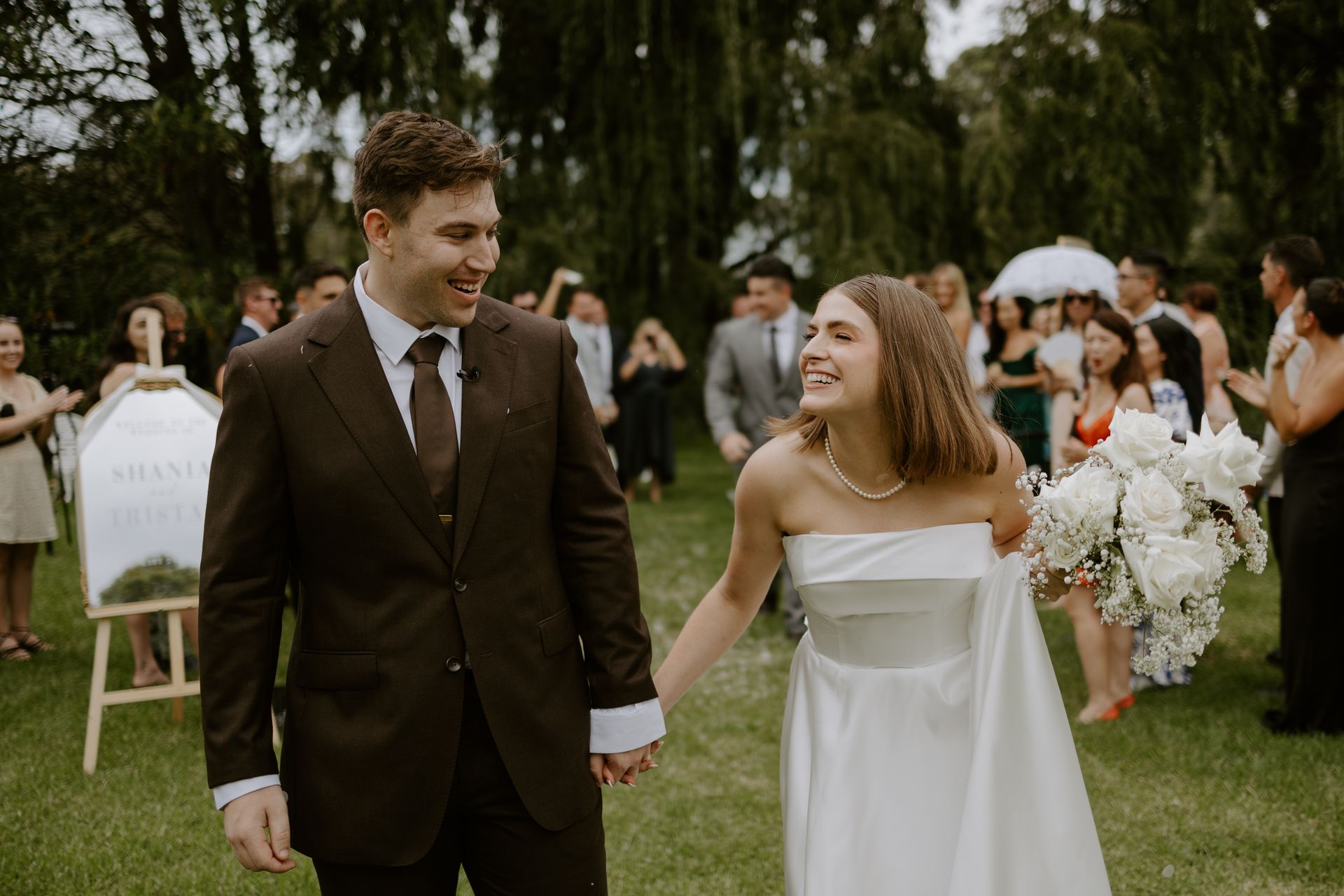 Newlyweds smiling, walking hand-in-hand through a grassy area, wedding guests in background.