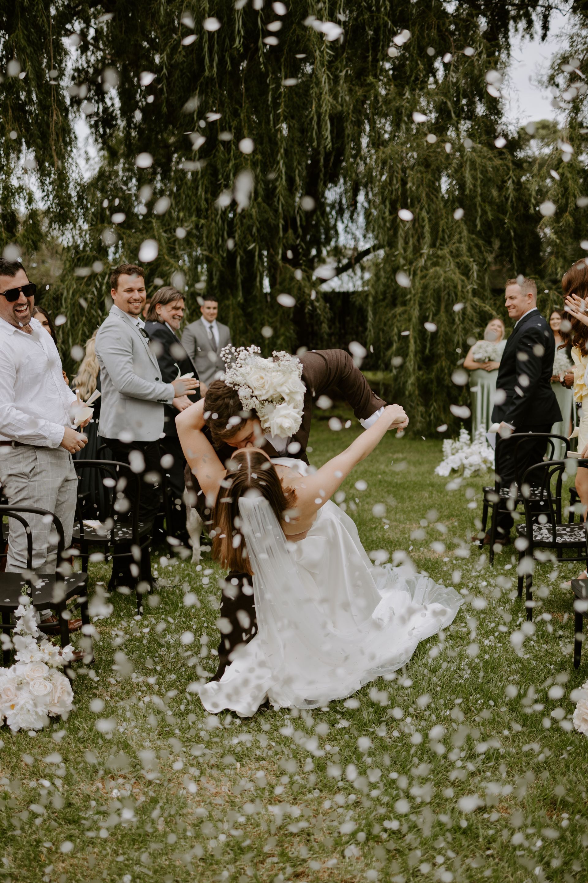 Newlyweds dip as confetti falls during outdoor wedding ceremony. Guests watch on the lawn.
