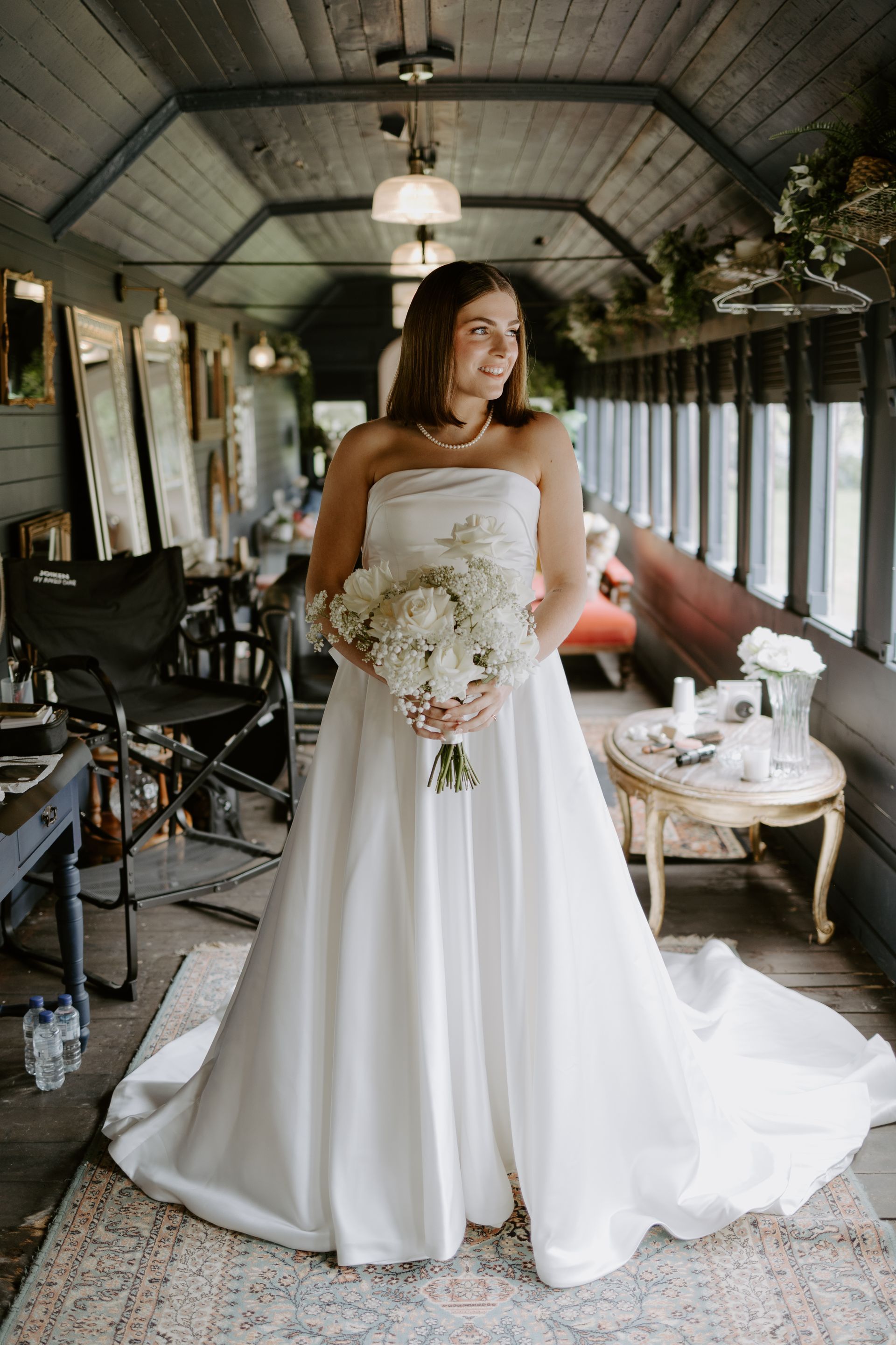 Bride in strapless white gown, holding a bouquet. Standing in an interior room with mirrors and decor.