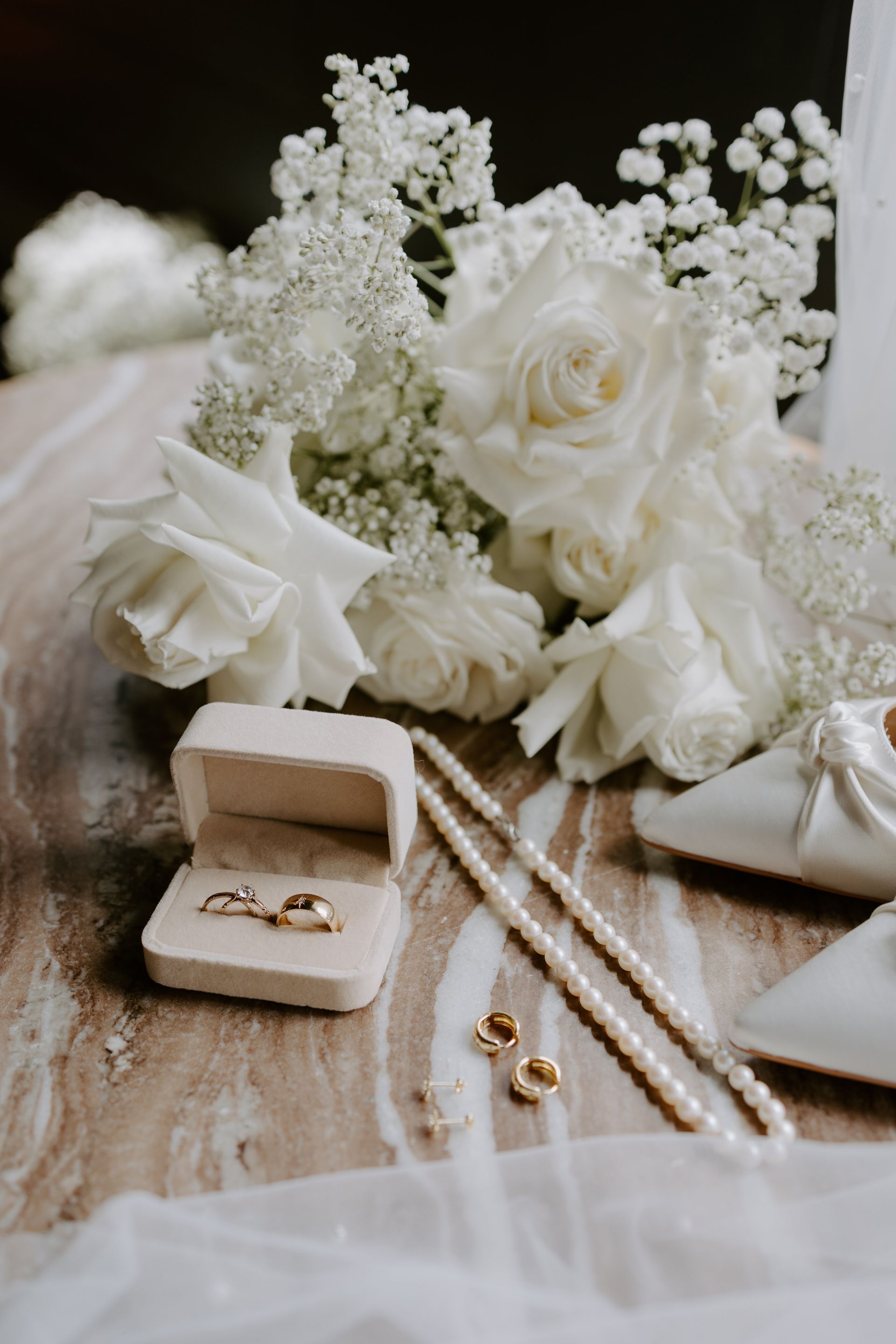 Wedding rings in a box, pearls, white flowers, and shoes arranged on a table.