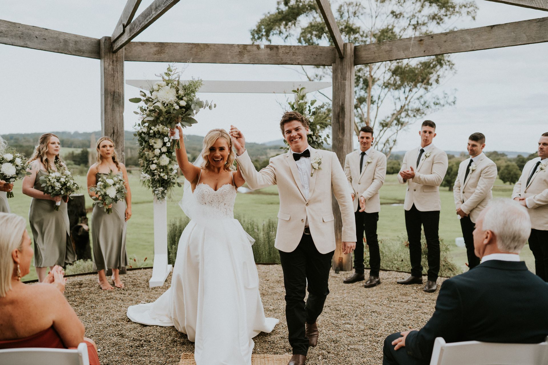 Newlyweds cheering, walking down the aisle after ceremony. Bride in white dress, groom in beige jacket.