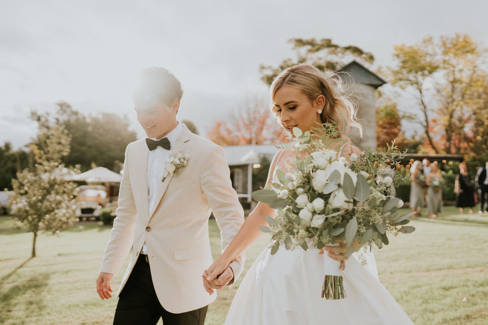 Bride and groom walking hand-in-hand in a sunny outdoor setting, the bride holding a bouquet.