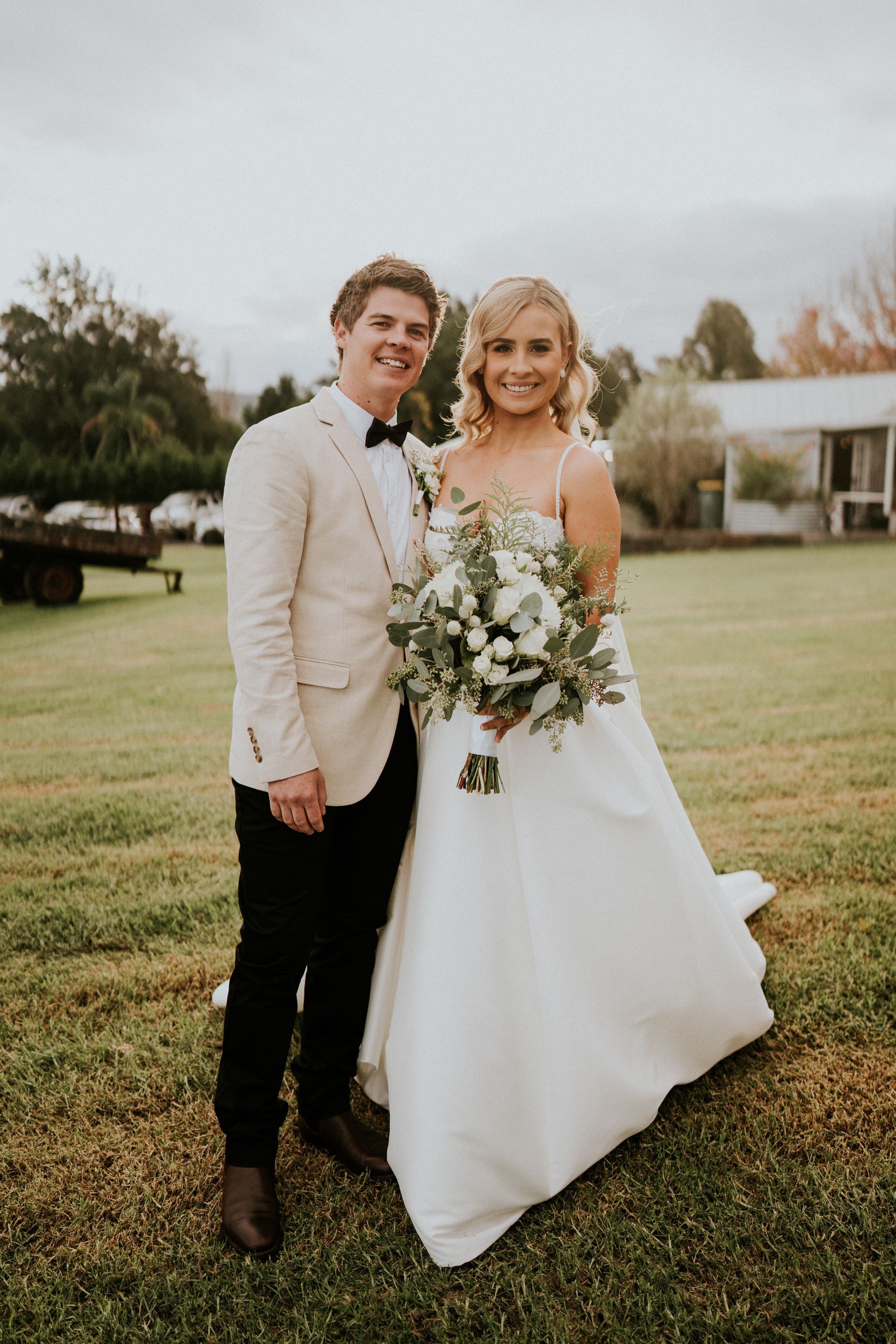 Bride and groom smiling, holding hands, standing on grass. Bride in white dress, groom in tan jacket.