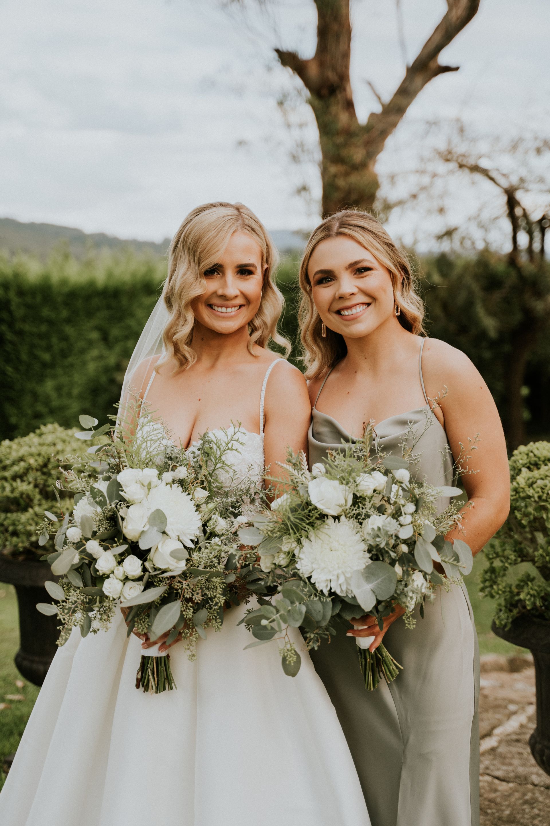 Bride and bridesmaid smiling, holding bouquets, in outdoor setting, trees in background.