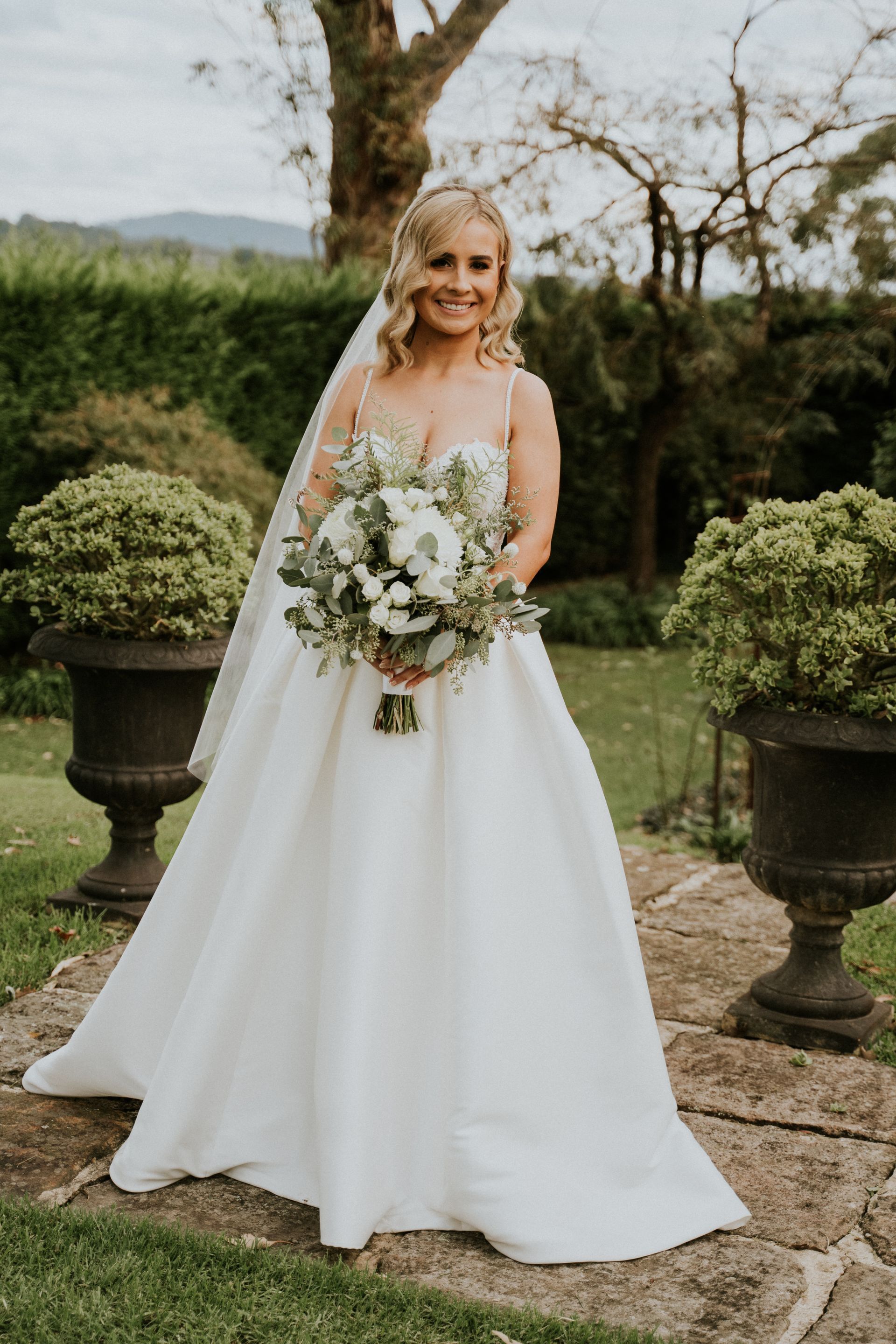 Bride in a white satin gown, holding a bouquet, standing outside.