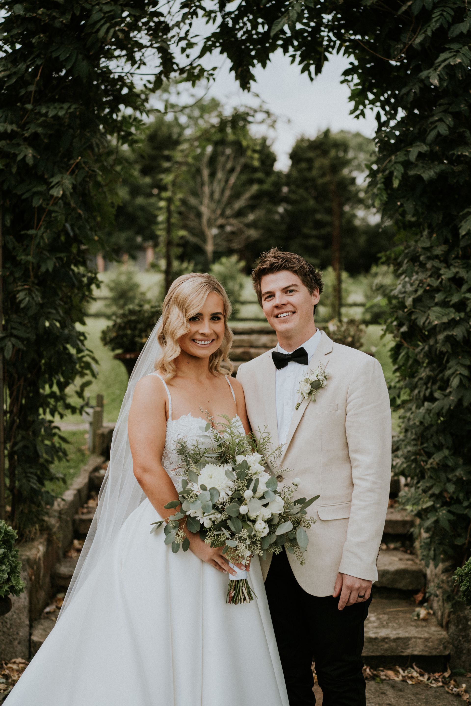 Bride and groom pose on stone steps beneath an ivy-covered archway. The bride holds a bouquet.