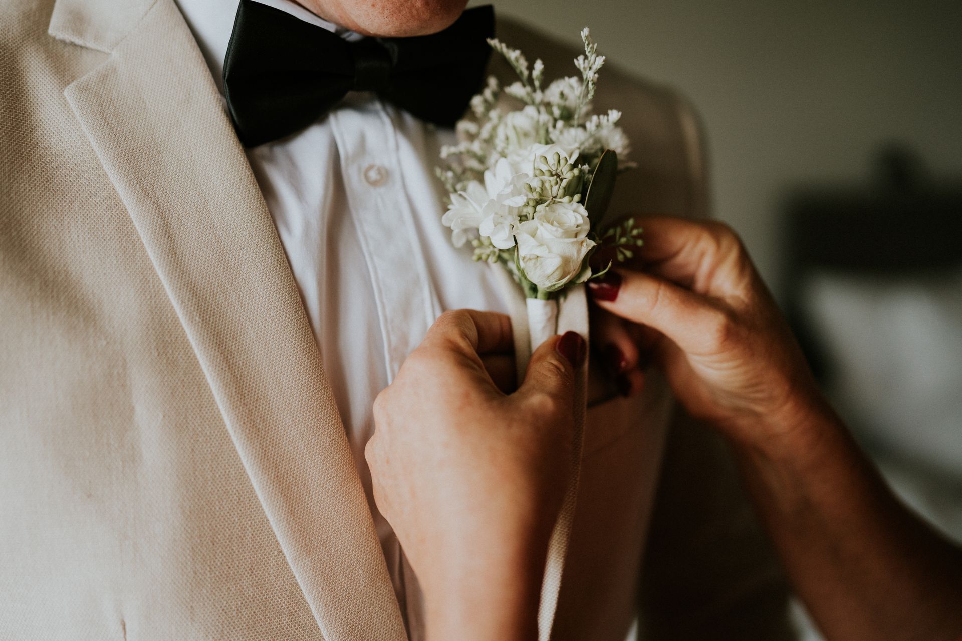 Woman pinning boutonniere on a man's lapel. He wears a beige jacket, white shirt, and black bow tie.