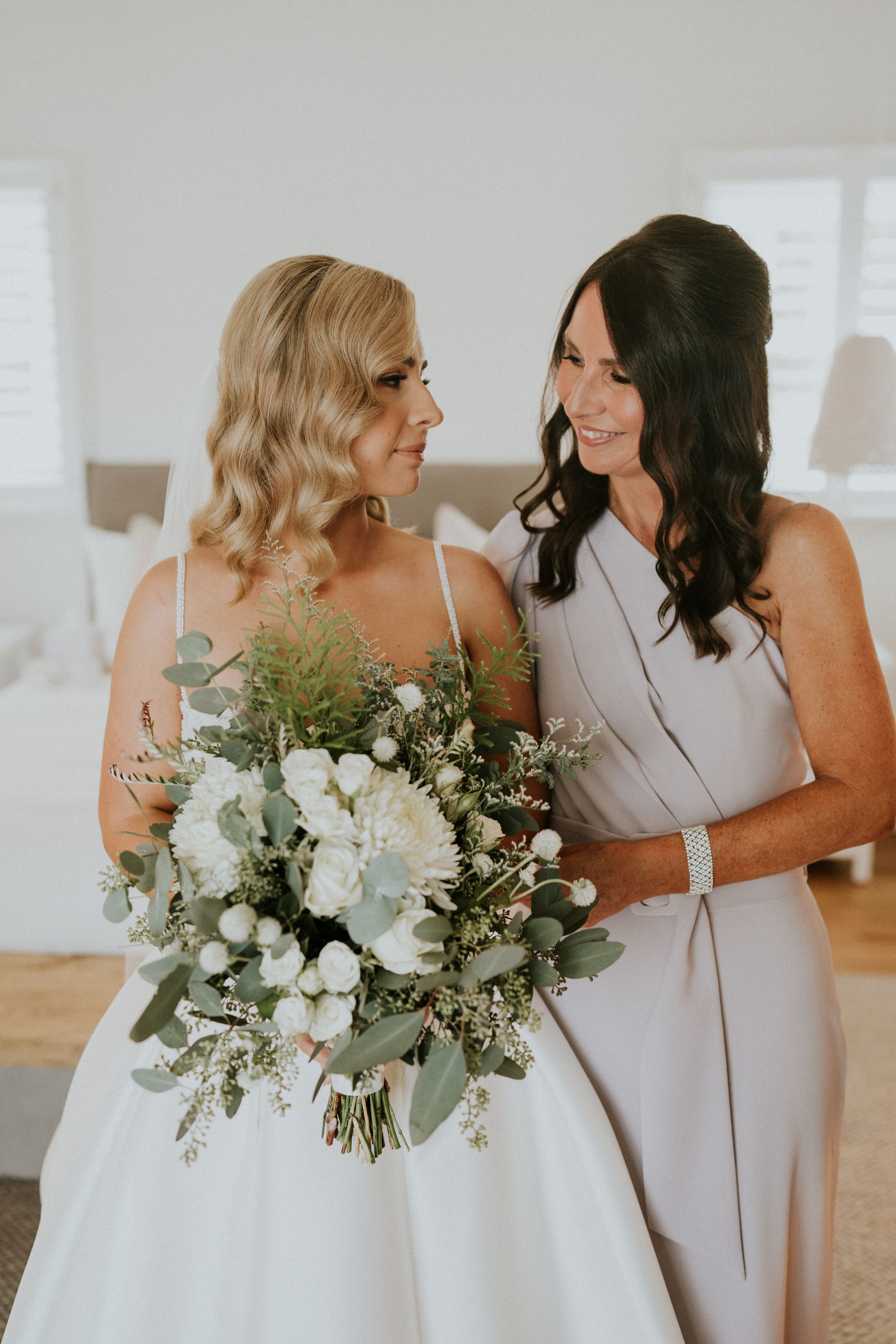 Bride holding bouquet smiles at woman in a lilac dress; indoor setting.