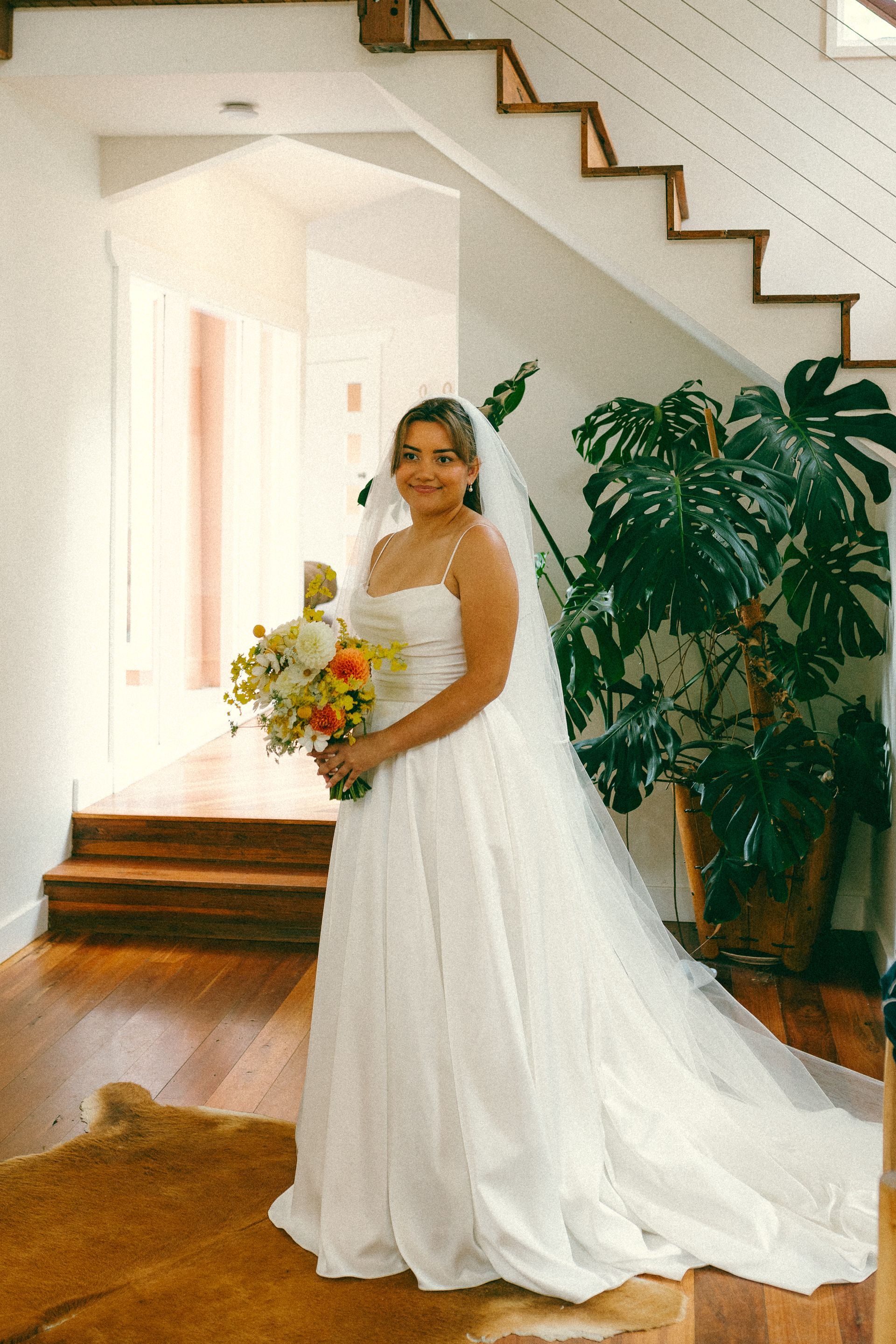 Bride in a white gown, holding flowers, standing near a staircase and large plant.