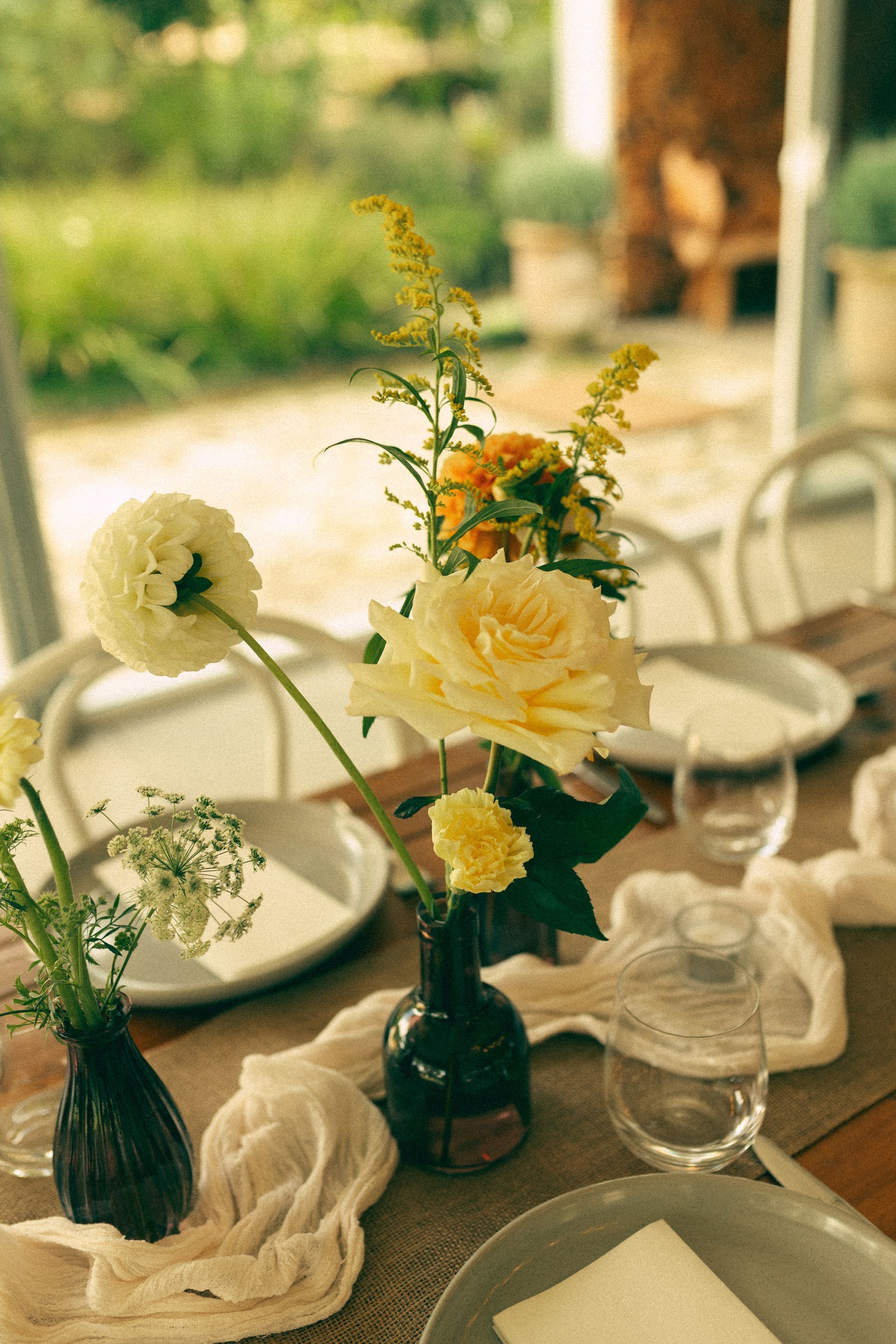 Floral centerpiece on a table set for a meal; yellow roses, white plates, glass cups, outdoor setting.