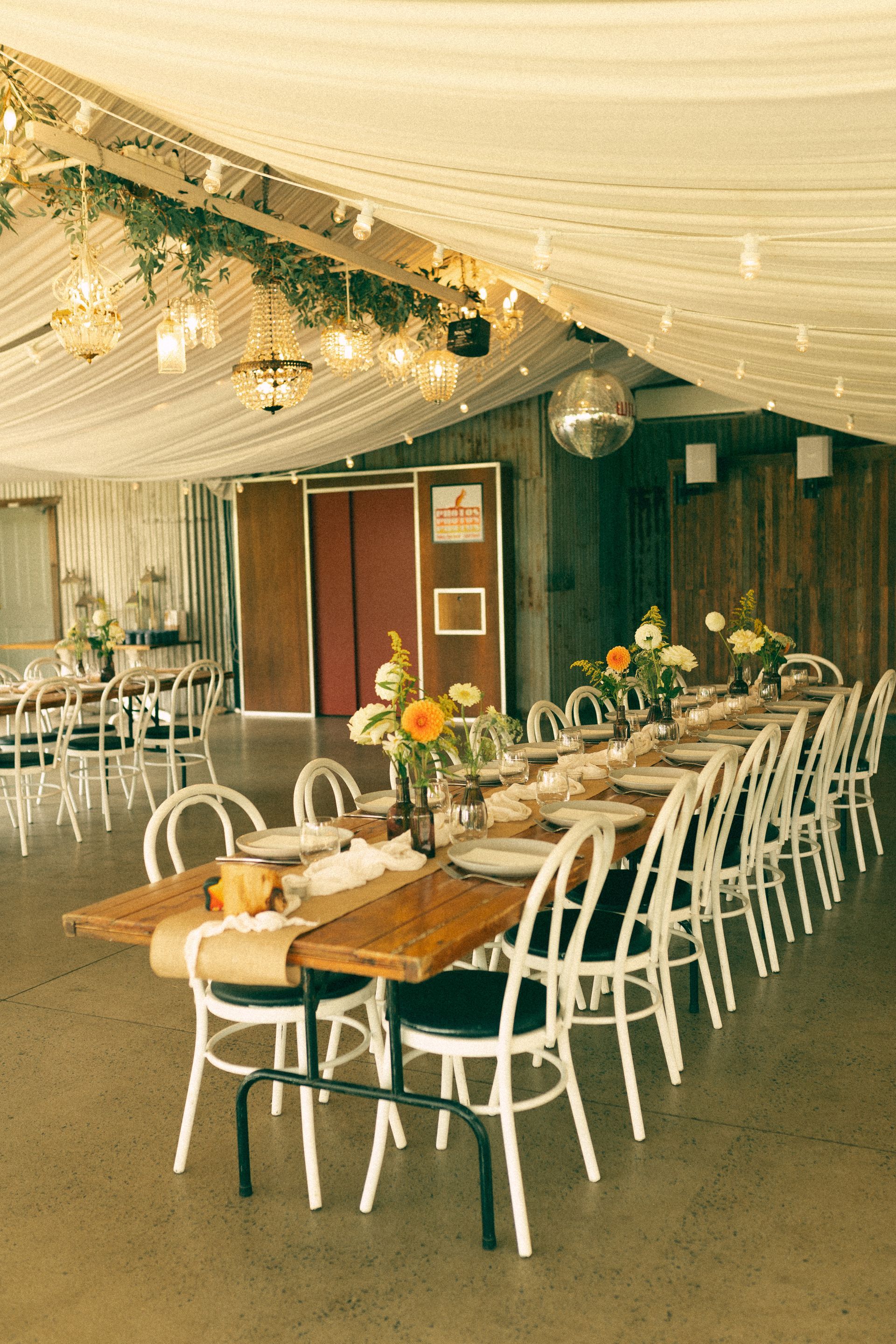 Long wooden table set for a party; white chairs; floral arrangement; string lights in a rustic setting.