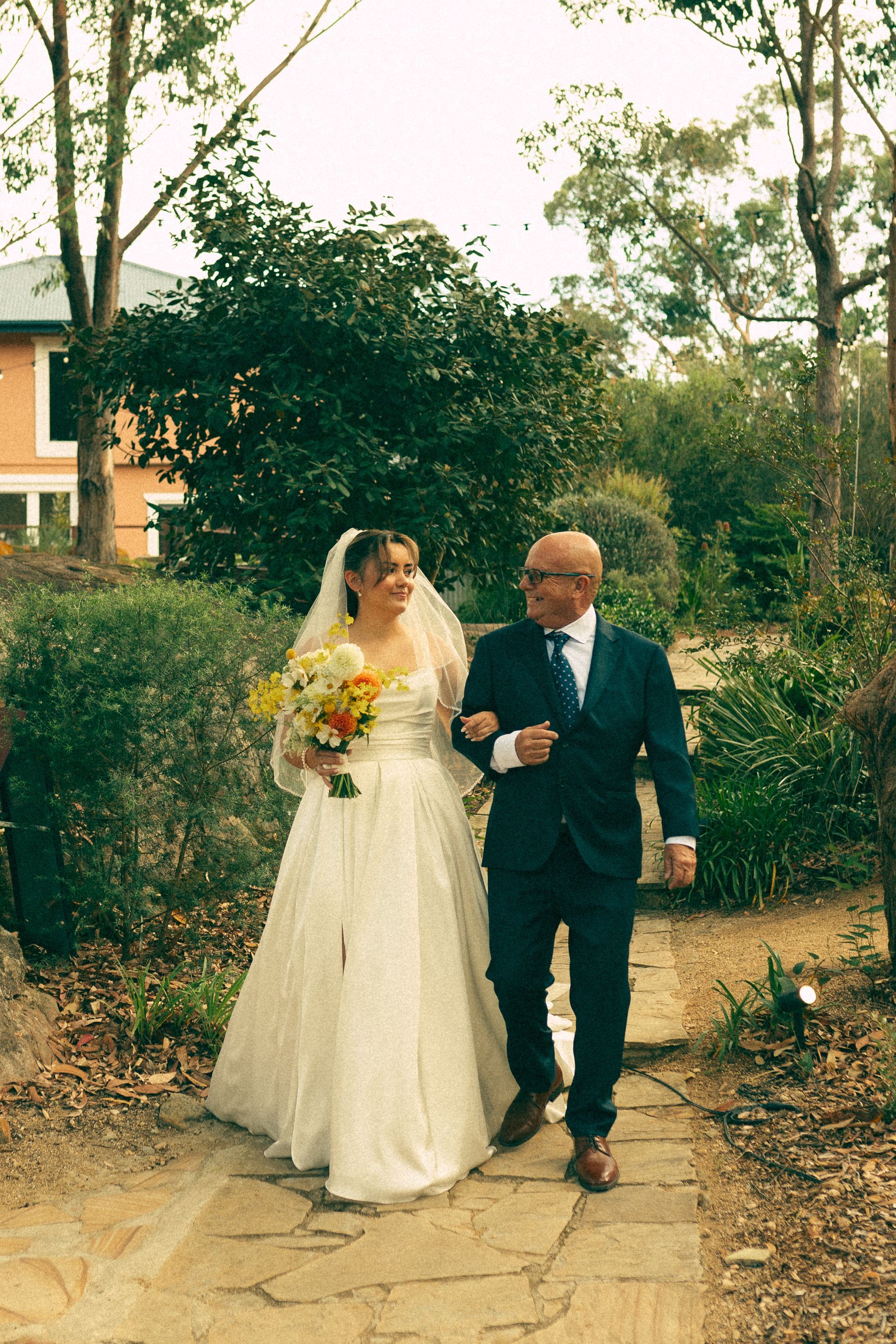 Bride in white dress walks with her father, arm in arm, down a stone path in a garden.