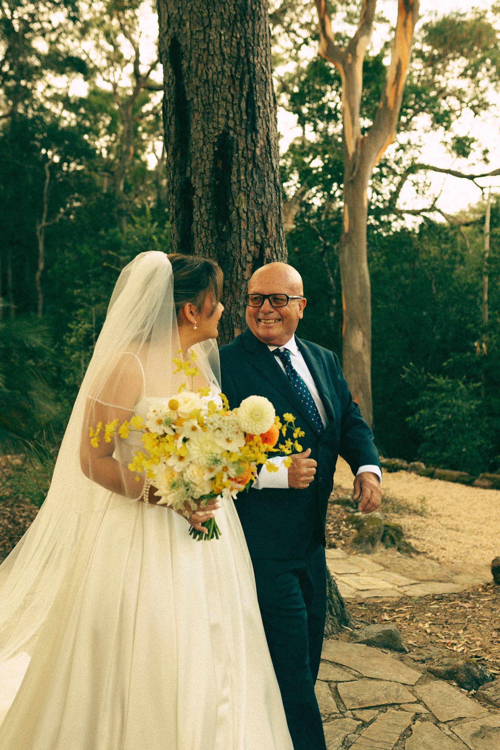 Bride in white gown with father, walking together in a park, smiling.