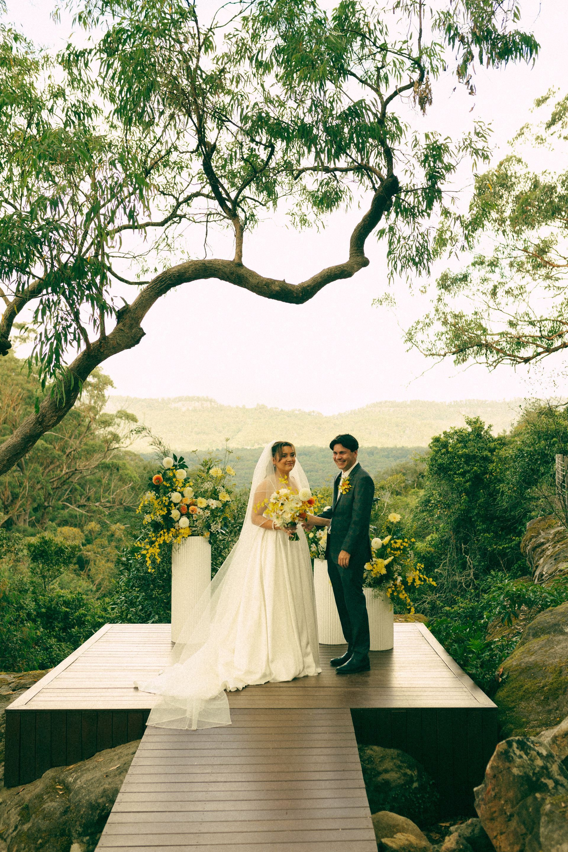 Bride and groom at outdoor wedding. Bride in white gown, holding flowers, groom in suit. Natural setting, wooden platform.